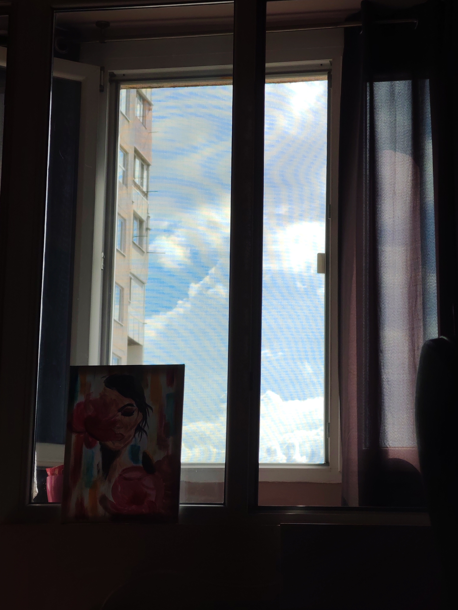 The image shows an indoor scene looking out a window. The window is divided into two sections, with a mesh screen visible in the right section. Outside the window, a building with multiple stories and windows is visible against a blue sky with white clouds. A dark curtain hangs on the right side of the window. In the foreground, on the windowsill, is a framed painting of a woman's face with abstract colorful brushstrokes. The overall lighting suggests it is daytime, with the sun likely behind the clouds, creating a slightly diffused light. The setting appears to be an apartment or residential building.