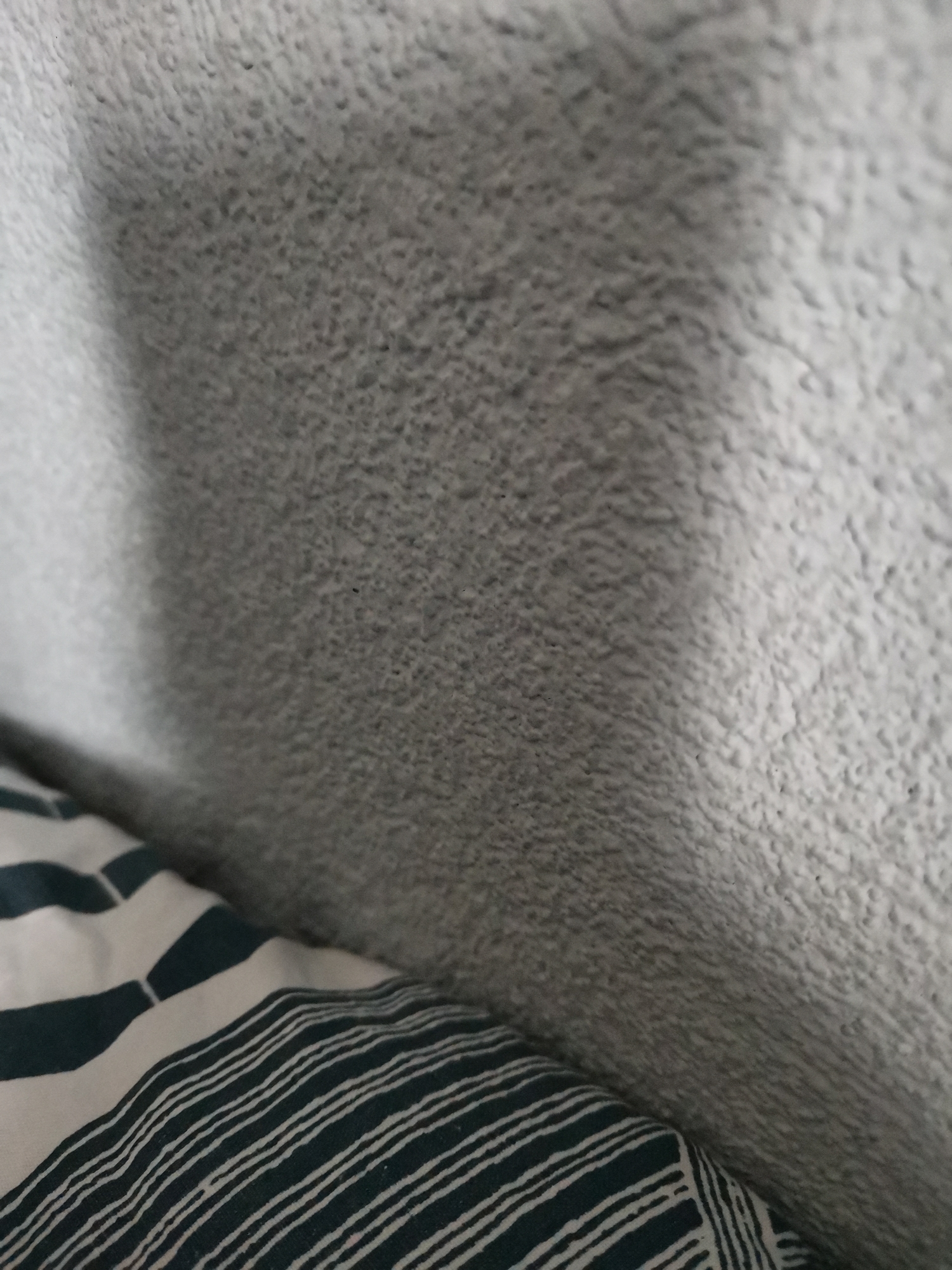 The image shows a close-up of a textured, light gray surface, possibly a wall or fabric. In the lower left corner is a striped textile, white and dark green or blue. The stripes are diagonal and parallel. The lighting is soft and diffuse. It's not possible to determine the location as Pokrovnik, Bulgaria, based on the given information. There are no people, actions, or interactions visible.