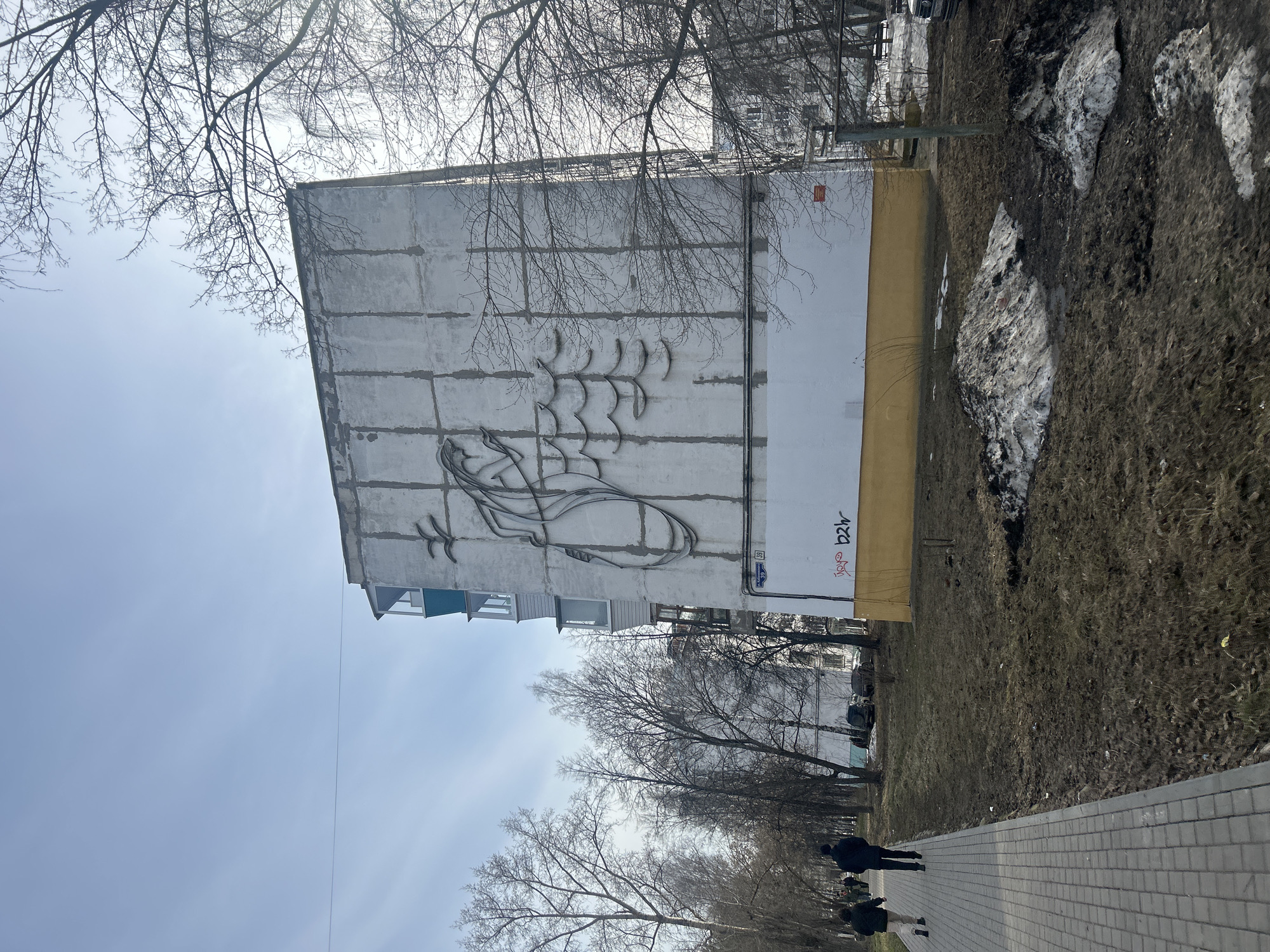 The image shows a five-story building in Chaikovsky, Russia, with a large abstract mural on its side, featuring a stylized image in black lines. The building appears to be made of concrete panels. The base of the building is painted in a yellow-beige color and there is some graffiti visible. A sidewalk runs along the lower part of the frame. In the background, a row of bare trees separates the sidewalk from a grassy area with some patches of snow. A few people are walking along the sidewalk. The sky is overcast.