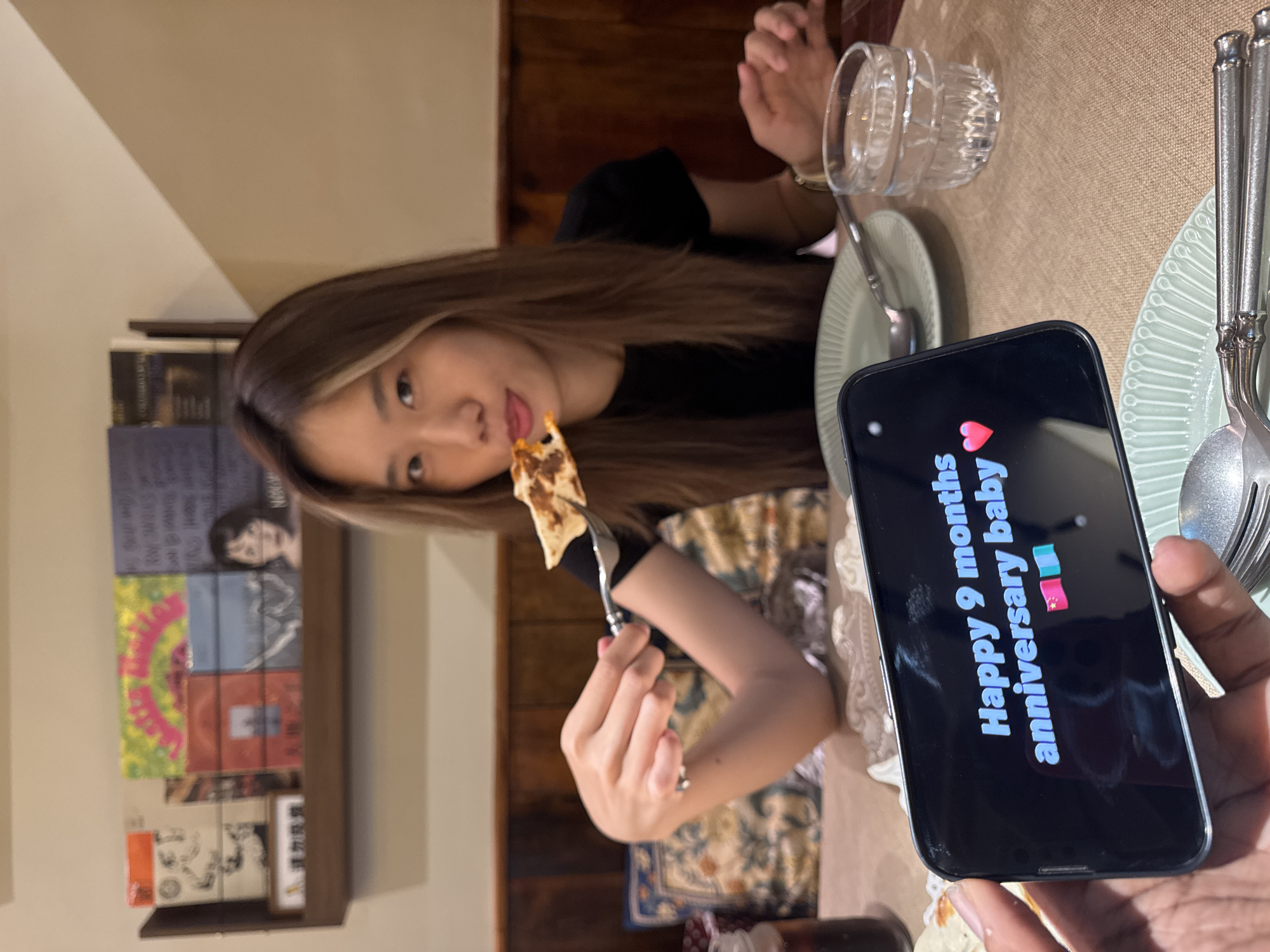 The image captures a celebratory moment at a dining table. A young Asian woman is seen holding a piece of food on a fork, poised to eat. A hand holds up a smartphone, displaying the words "Happy 9 months anniversary baby" with heart and gift emojis. The dining table is set with plates, cutlery, and a glass. In the background, a wooden wall paneling and a shelf holding stacked books or records are visible. The scene suggests an intimate, personal occasion being documented with a mobile device. The location cannot be identified as Yuzhong District, China, based on the visual elements in the image.