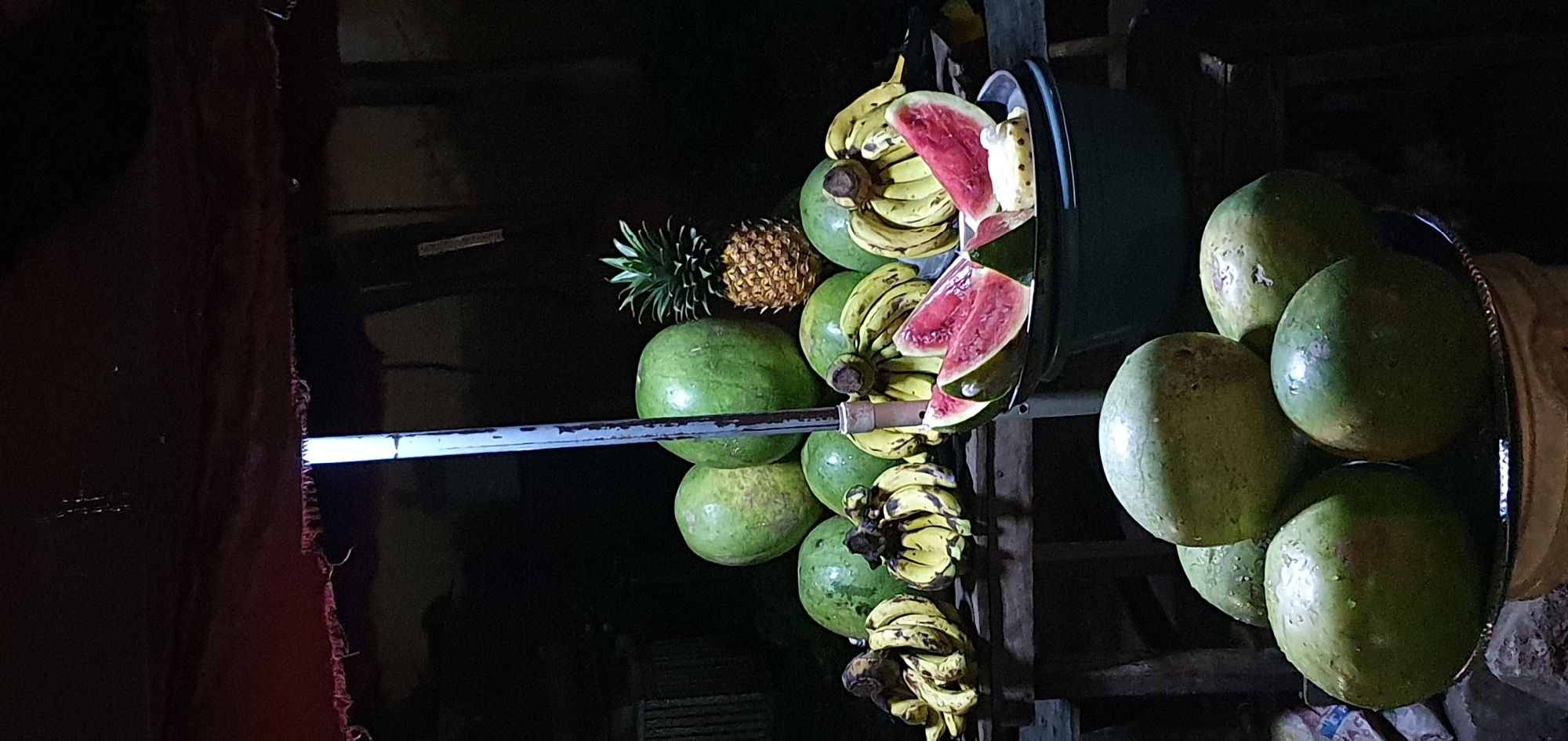 A nighttime outdoor stall displays a variety of fruits in Bukuru, Nigeria. A dark red tarp is stretched overhead, with a vertical light pole running down the center.
In the background, a variety of large, round, green fruits, possibly watermelons or gourds, are stacked. To their right, a spiky pineapple sits atop one of the green fruits. In front of these, a collection of bananas, some with brown spots, are arranged on a wooden surface.
In the foreground, a dark green bowl holds several slices of bright pink watermelon, revealing their seeds. To the left, another stack of large, round, green fruits sits on a silver platter, which is itself resting on a woven base. The overall lighting is dim, with the fruits illuminated by the light pole and potentially other unseen light sources, creating strong shadows. The scene appears to be a food stall or market stall.