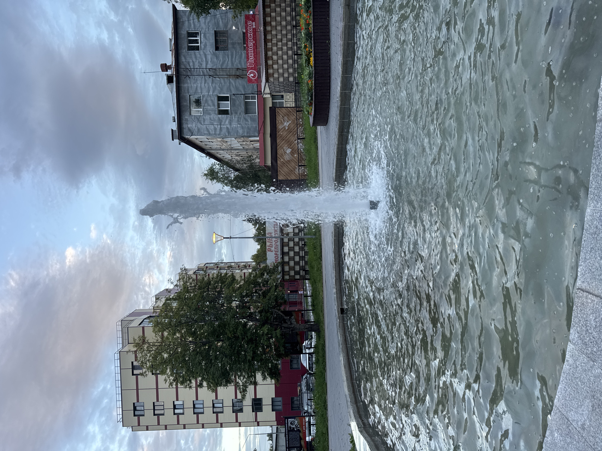 The image shows a fountain with water jets spraying upwards in the foreground. In the background, there are several buildings. On the left is a modern, multi-story building with a beige and red facade. To its right, a large green tree is partially obscuring it. In the center background, a street lamp is visible. Further to the right, there is a grey, older building with multiple windows. A red sign with white text and a circular logo is attached to the side of this building, with the text "Пионерское" visible. Between the buildings, there are signs and structures, including a brown fence with a decorative pattern. The sky is overcast with a mix of grey and pale blue clouds. The overall impression is of an urban setting, likely a public space with a fountain and commercial or residential buildings. This scene is located in Yelizovo, Russia.