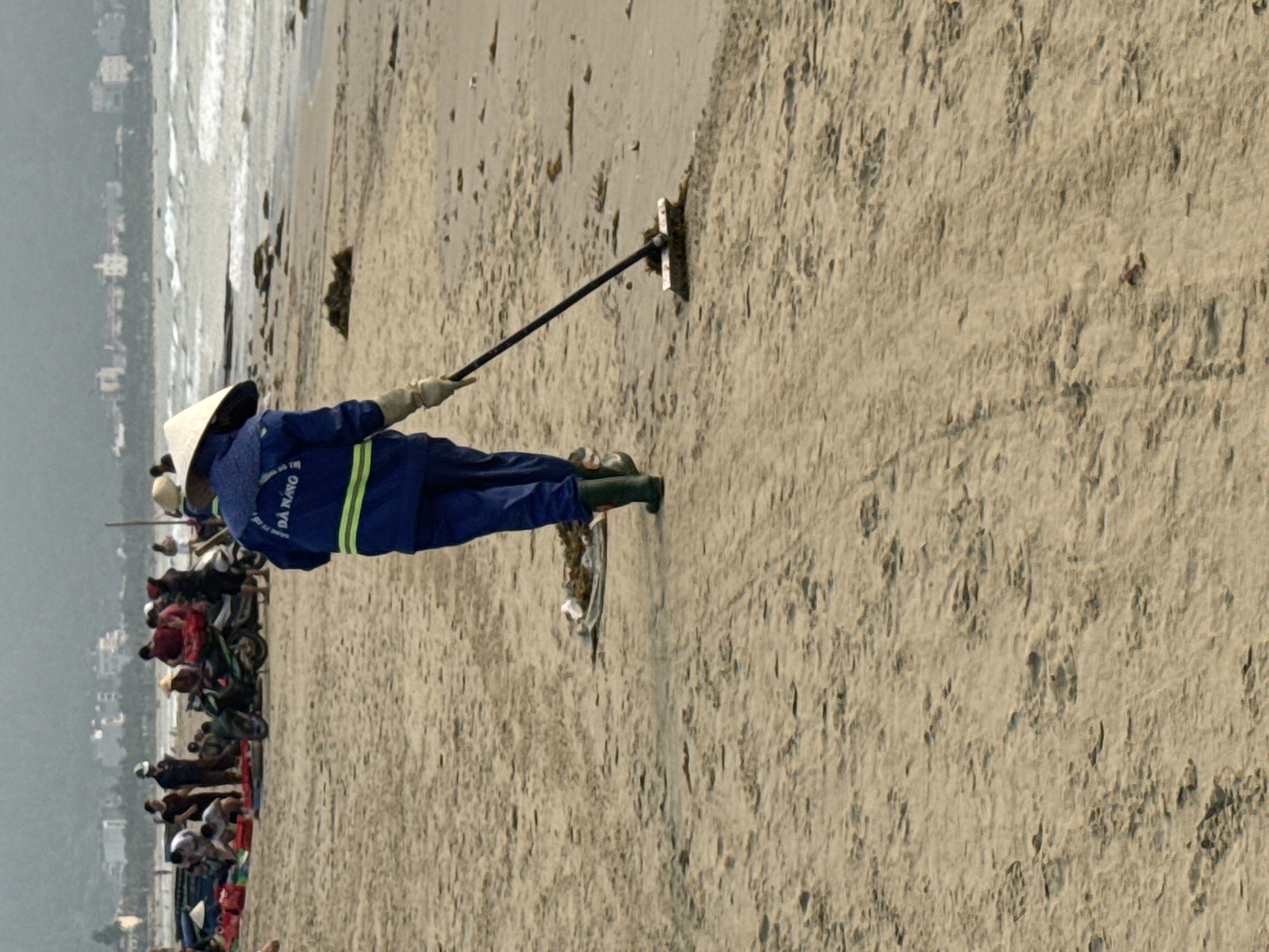 A beach scene in Đà Nẵng, Vietnam. A person wearing a conical hat, blue uniform with green stripes, and rubber boots is cleaning the sand with a long-handled tool. A group of people and parked scooters are visible along the beach behind the cleaner. The ocean and cityscape are visible in the distance. The sand appears to be slightly damp and marked with lines from the cleaning tool.
