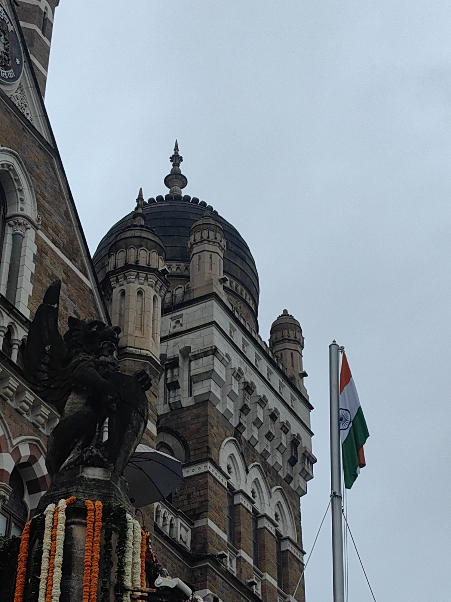 The image shows a low-angle, exterior shot of the Chhatrapati Shivaji Maharaj Terminus (CSMT) in Mumbai, India. The building, constructed in Victorian Gothic Revival architecture, features brown brickwork, white stone detailing, and a prominent dark dome topped with spires. A large, winged stone sculpture, possibly a lion or griffin, stands to the left of the frame, adorned with garlands of orange and white flowers. In the background, the Indian flag flies from a flagpole against a grey, overcast sky. The flag's vibrant colors – saffron, white, and green, with the Ashoka Chakra in the center – are clearly visible. The overall atmosphere is solemn and patriotic.