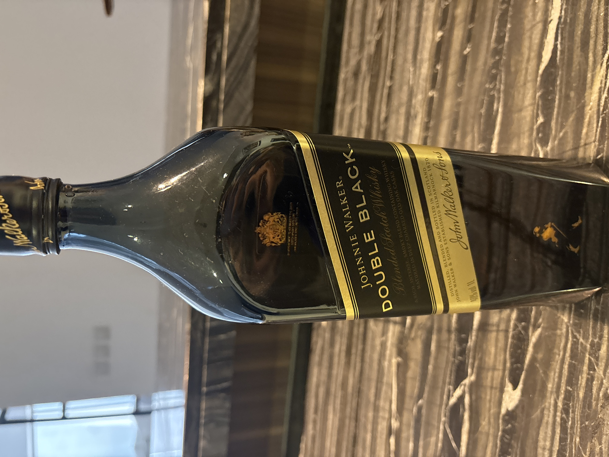 A tall, dark, rectangular bottle of Johnnie Walker Double Black Blended Scotch Whisky stands upright on a polished, marble-patterned surface. The bottle is mostly black with gold lettering and accents. The label prominently displays "JOHNNIE WALKER" and "DOUBLE BLACK Blended Scotch Whisky." Below this, smaller text describes the blend and maturation. The top of the bottle has a black cap with gold lettering. The background is blurred, showing what appears to be a modern interior with a large window on the left, suggesting an indoor setting in Dubai, United Arab Emirates. There are no people visible in the image.