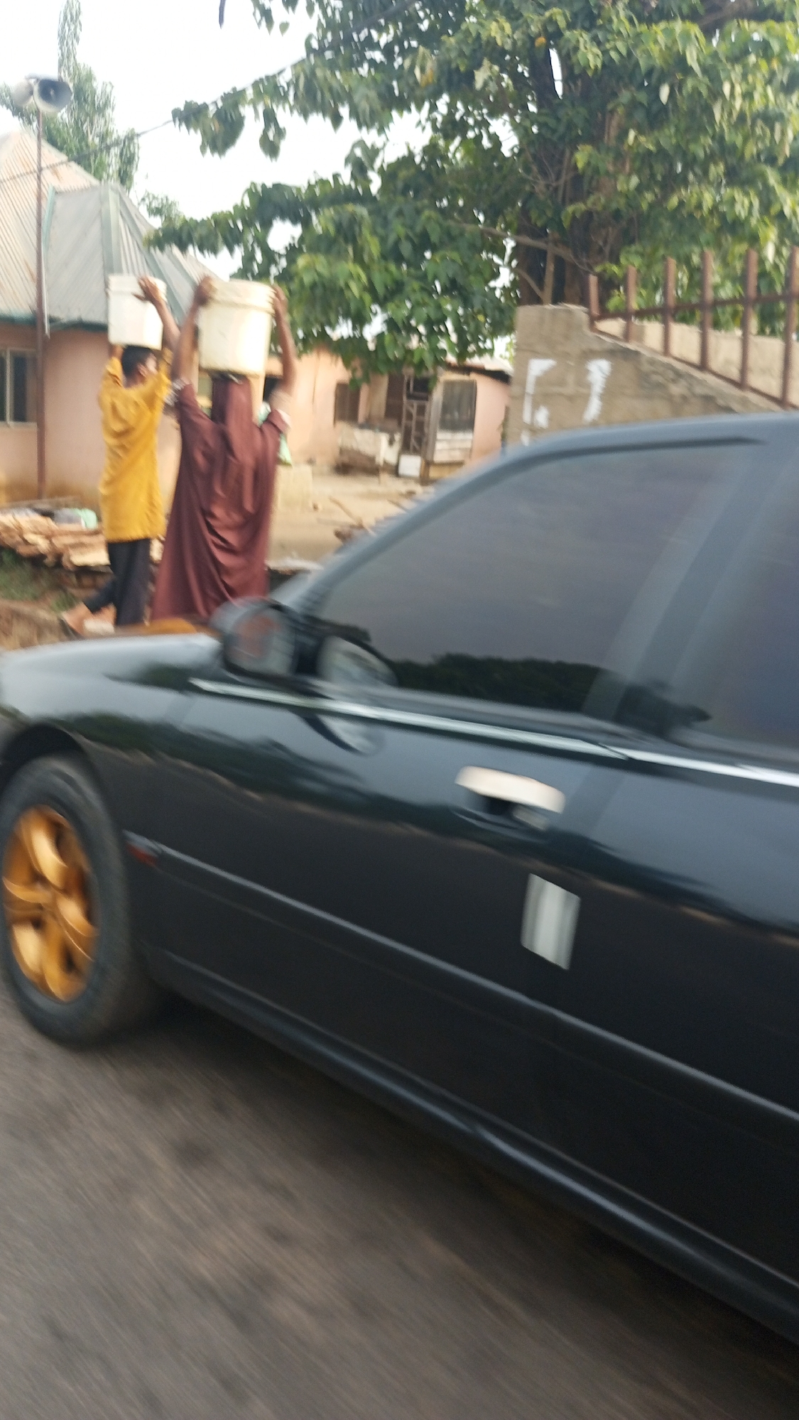 This image depicts a street scene in Zango-ndaji, Nigeria, capturing a blend of traditional activity and modern elements. In the mid-ground, two individuals, likely women, are prominently featured, walking and carrying large white buckets expertly balanced on their heads. The person on the left wears a bright yellow tunic over dark trousers, while the one on the right is dressed in a dark brown, flowing garment. Their faces are indistinct due to motion blur and distance.
Behind the people, a residential area is visible, characterized by simple houses with light-colored walls and corrugated metal roofs. One house on the left features a distinct pole with an attached loudspeaker or siren. Lush green trees with dense foliage are scattered across the background, providing shade and indicating a natural environment. A concrete wall or fence is also visible towards the right side of the background.
The foreground and right side of the image are dominated by a black sedan car. The car features dark tinted windows and striking gold-colored rims on its front wheel, suggesting a personalized touch. Its body reflects the ambient light, and a chrome or silver door handle is visible. The road surface below the car appears textured and blurred, consistent with either a moving vehicle or a quick snapshot.
The lighting suggests it is daytime, possibly late morning or early afternoon, with a bright, somewhat overcast sky. The overall scene conveys a snapshot of daily life, where traditional practices like head-carrying coexist with contemporary vehicles. No visible text is discernible in the image.