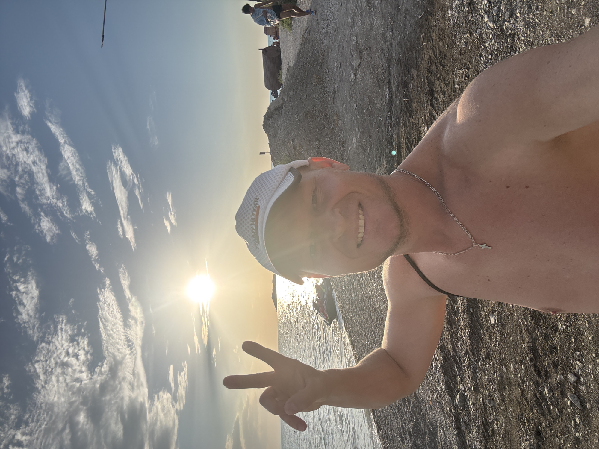 A man in a white baseball cap and a silver cross necklace is taking a selfie on a beach. He is shirtless and smiling, holding up two fingers in a peace sign. The sun is setting over the ocean, casting a warm glow on the scene. In the background, a jet ski is parked on the shore, and a few people are walking along the beach. The beach is made up of dark pebbles and sand. The location is not Zharbulak, Kazakhstan, as this is a coastal scene.