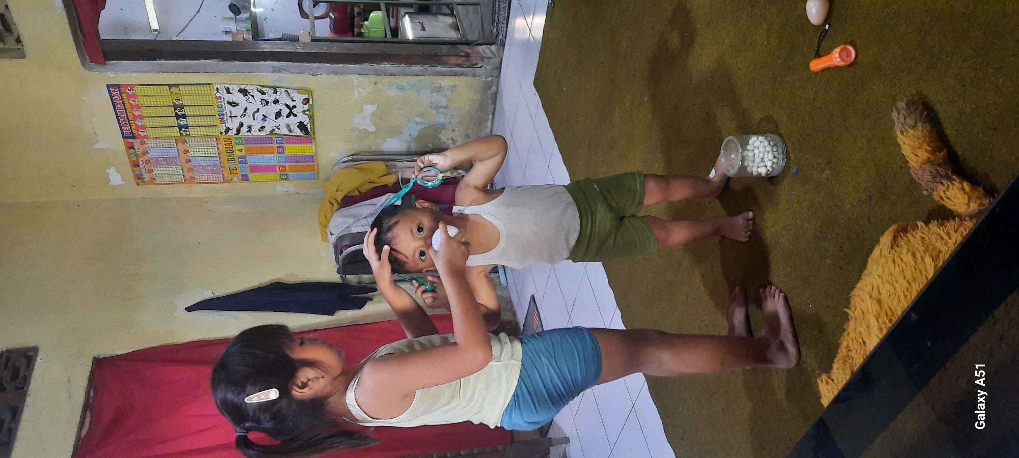 In a room in Ploso, Indonesia, a young girl with her hair in a ponytail, wearing a light-colored tank top and blue shorts, is attempting to cut the hair of a young boy. The boy, wearing a white tank top and green shorts, has a white object, possibly a roll of tissue, placed over his nose and mouth. He is holding a pair of green scissors in his right hand. Both children are barefoot and standing on a green rug. A clear glass jar filled with white pills or capsules sits on the rug near the boy's feet. To the right, an orange flashlight and a white oval object are also on the rug. In the background, a poster with numbers and images of insects is displayed on a light-colored wall. A red curtain hangs to the left, and clothes are draped over a rack behind the children. The overall impression is a casual, indoor domestic scene.