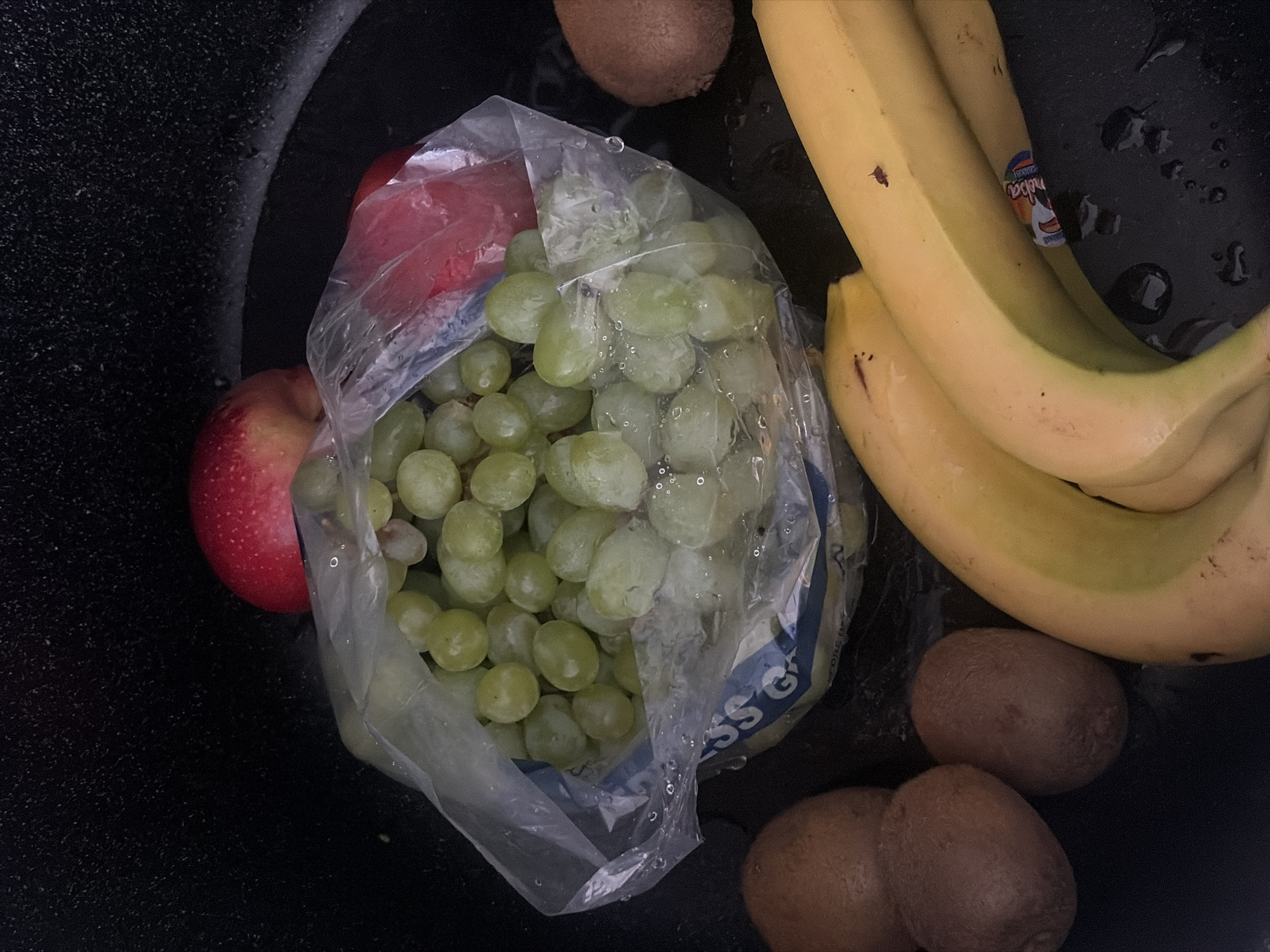 A top-down view reveals a black container filled with various fruits. In the center, a plastic bag contains green grapes. An apple, partially covered by the bag, is positioned to the left. A bunch of bananas is situated to the right. Three kiwis are located at the bottom right, with an additional kiwi near the top. There is no indication of this photo being taken in Saint Petersburg, Russia.