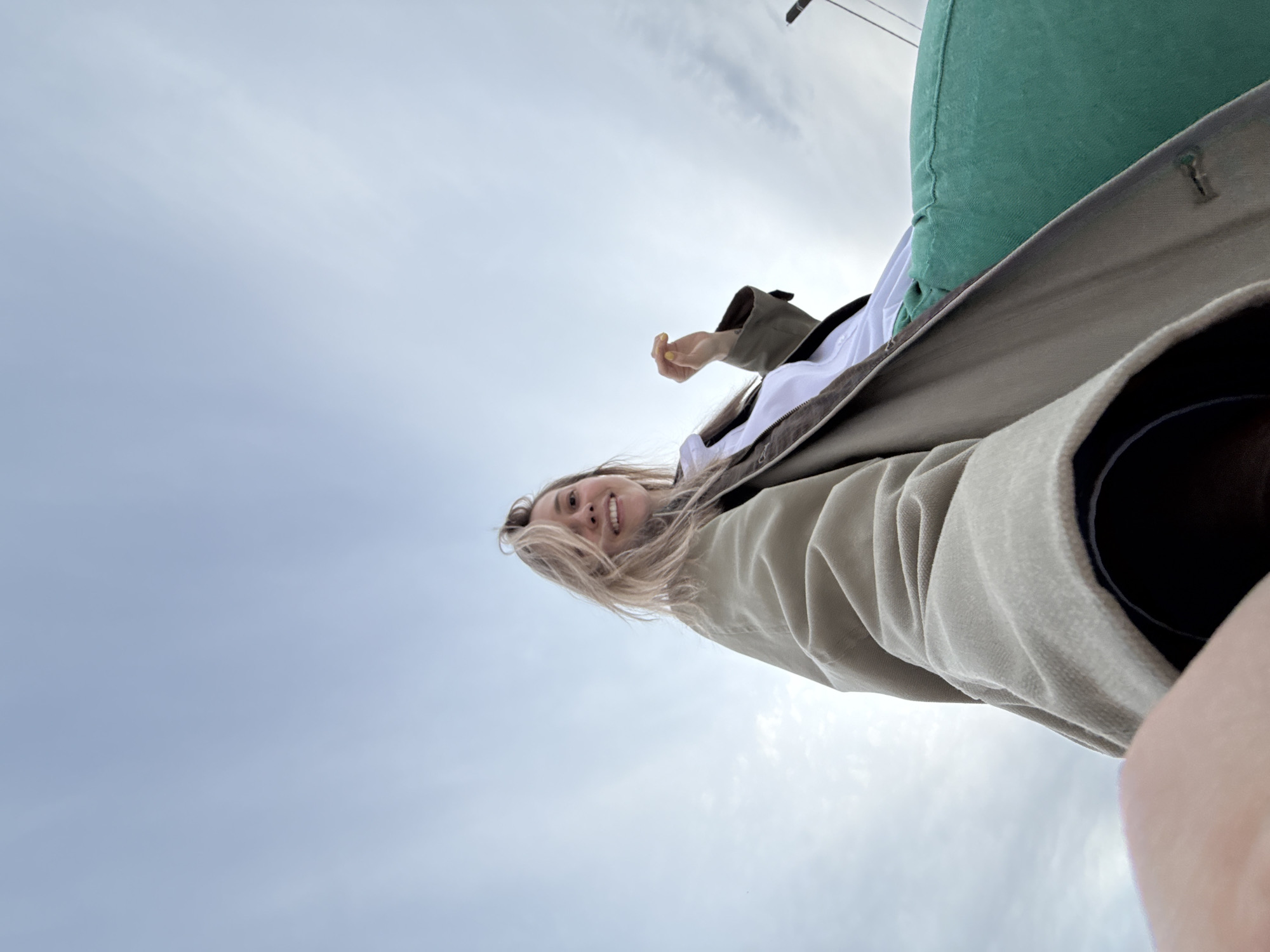 A low-angle shot shows a woman with blond hair and a white shirt under a tan coat smiling at the camera. The background is a cloudy sky, and a green bag is visible in the upper right corner. A dark object, possibly a camera lens, is visible in the lower right corner, suggesting the woman took the photo herself. The location is unknown, but it is requested to be Chaikovsky, Russia. Without further information, it cannot be confirmed.