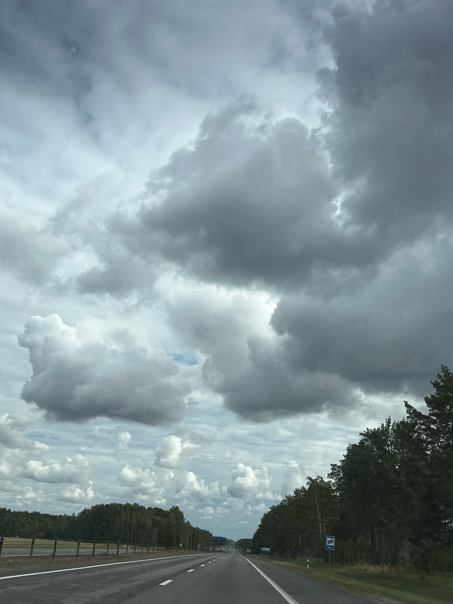 The image depicts a highway scene under a cloudy sky, likely in the Вяззеўскі сельскі Савет, Belarus. The viewpoint is from a vehicle moving forward on a two-lane asphalt road. Trees line the right side of the road, and a fence runs along the left side, bordering a field with more trees in the distance. The sky is filled with large, gray and white cumulonimbus clouds, with some lighter patches indicating diffused sunlight. There are no people or vehicles visible in the immediate foreground or on the road. A blue sign with a camera icon and the number "800m" is visible on the right side of the road. The overall atmosphere is overcast and somewhat dramatic due to the cloud formations.