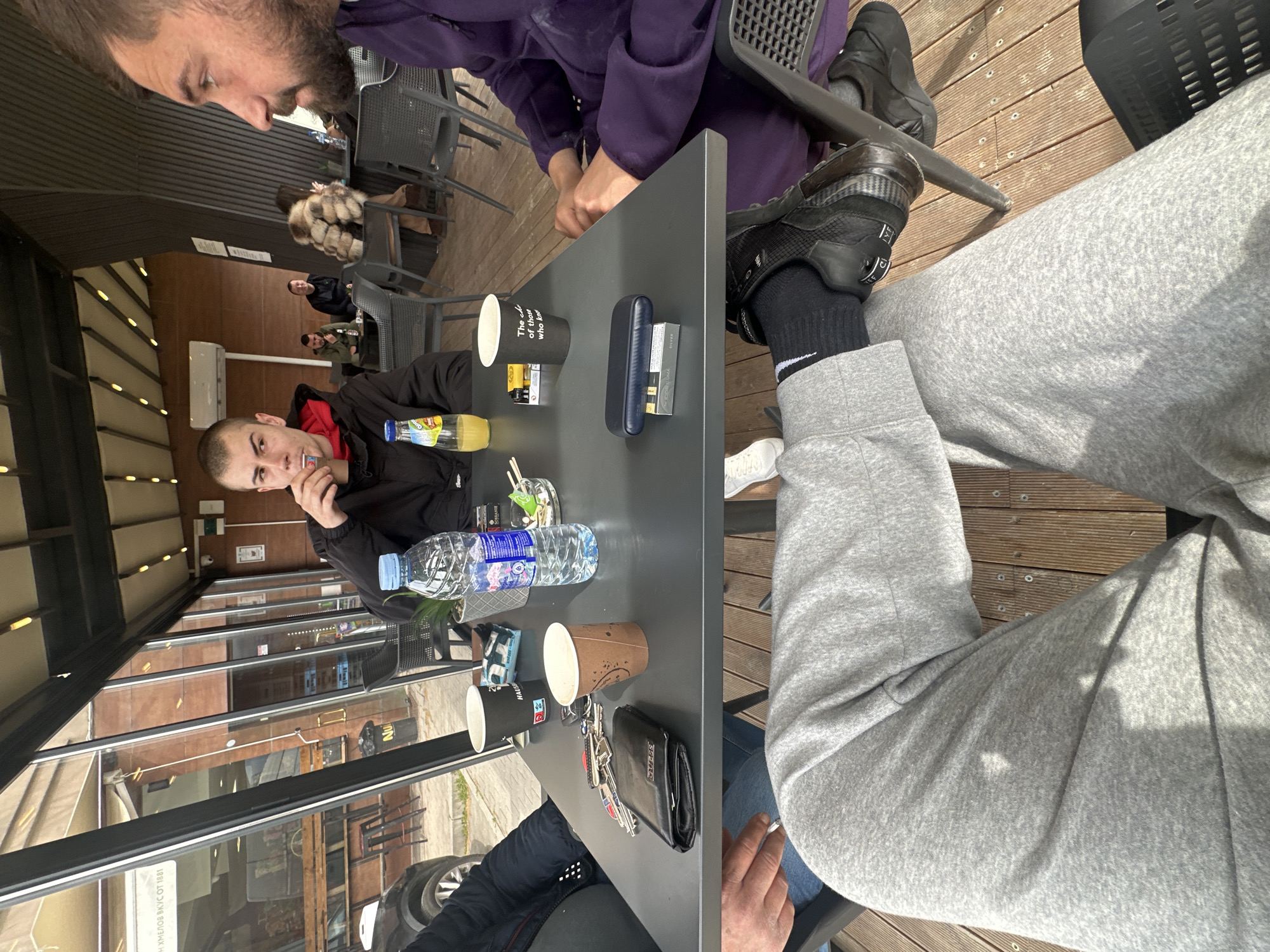 The image shows a group of people sitting at an outdoor cafe in Plovdiv, Bulgaria. They are seated at a gray metal table with cups, a water bottle, a wallet, keys, juice, cigarettes, and an electronic cigarette. Two men are visible on the far edges of the image. One is in a purple shirt and the other is in a black jacket. The environment is an outdoor cafe with a partially enclosed patio. The floor is wooden. Other people are seated at tables in the background.