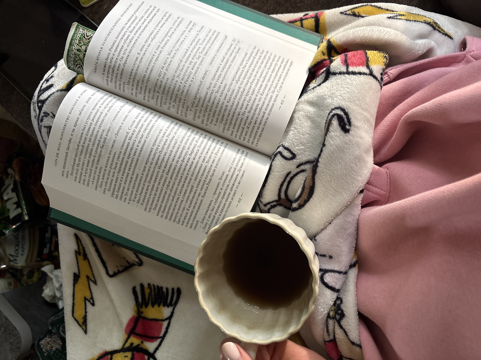 The image depicts a book in Russian, titled "САРА ДЖ. МААС. КОРОЛЕВСТВО КРЫЛЬЕВ И РУИН" (Sarah J. Maas, Kingdom of Wings and Ruins), lying open on a blanket with a Harry Potter-themed design. Adjacent to the book is a ceramic cup containing a dark liquid, presumably tea or coffee. A person's hand with a manicured nail is visible near the cup. The scene is set indoors with a pink garment and what appears to be a packaged item with the word "MOCKBA" (Moscow) visible in the background. No information is present to place the image in the city of Khimki, Russia.