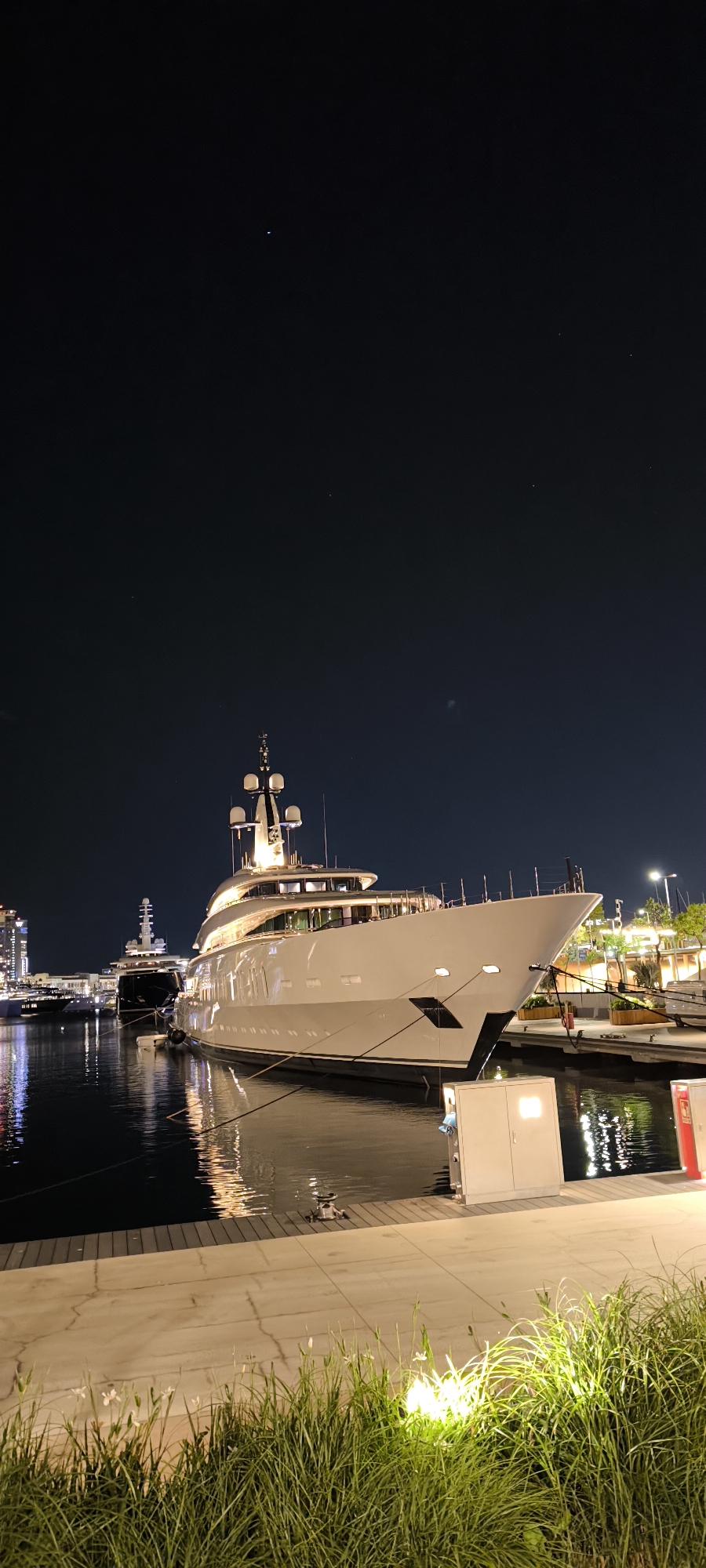A nighttime scene shows a large white yacht docked in a marina, likely in Barcelona, Spain, given the presence of modern architecture in the background. The yacht is illuminated, casting reflections on the water. Docking infrastructure, including power boxes and mooring equipment, is visible along the quay. The foreground features grass and paving stones. The sky is dark, with a few visible stars. No people are immediately visible in the scene. Other yachts are docked in the background.