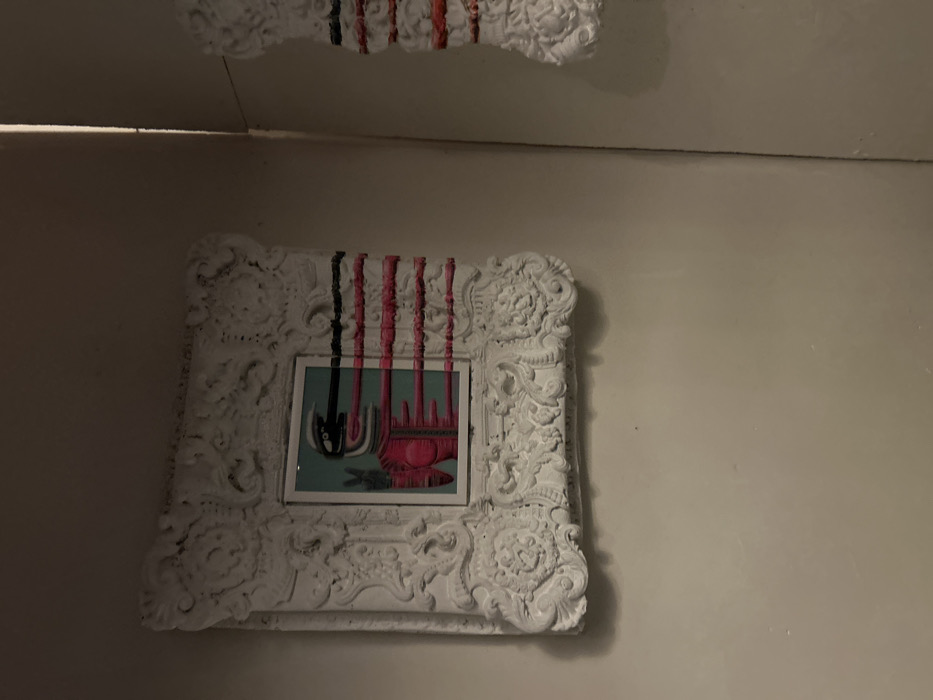 An ornate, white frame encases a colorful abstract painting. Black and pink painted lines descend from the top of the frame, partially obscuring the art within. The painting itself features a teal background with a red, ambiguous object dominating the lower portion. A black, cup-like form hangs suspended in the upper left of the frame. Above the frame, another decorative white piece with similar painted lines is partially visible against a plain white ceiling. The entire scene is set against a light beige wall.