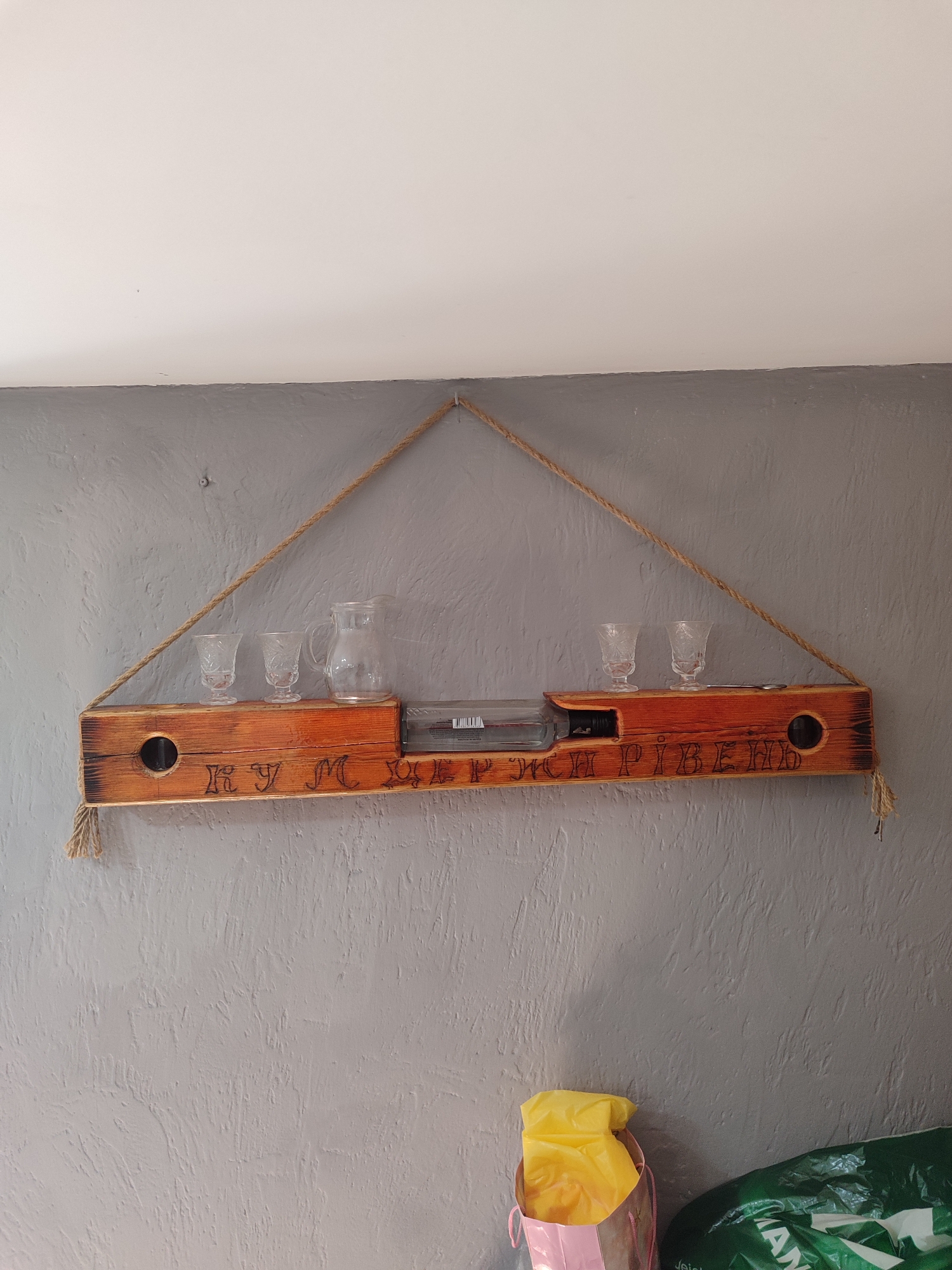 The image displays a wooden shelf with carved text, suspended by rope against a textured gray wall. On the shelf, there are several items including a glass pitcher, three small decorative glasses, and a partially visible bottle, possibly containing alcohol. The shelf itself appears to be made from rough-hewn wood, stained a warm orange-brown. The carved text on the shelf reads "RYM. RIVNE". There are also circular holes on either end of the shelf, possibly for decorative purposes or for inserting hanging materials. The rope used to suspend the shelf is thick and braided, with tassels hanging down from the ends. The wall behind the shelf is painted a matte gray with a visible texture. In the lower right portion of the image, a pink shopping bag with yellow contents and a large green plastic bag with visible white text are present, suggesting a domestic setting. The lighting in the image is even, indicating an indoor environment. The overall impression is of a rustic and perhaps homemade decorative item.