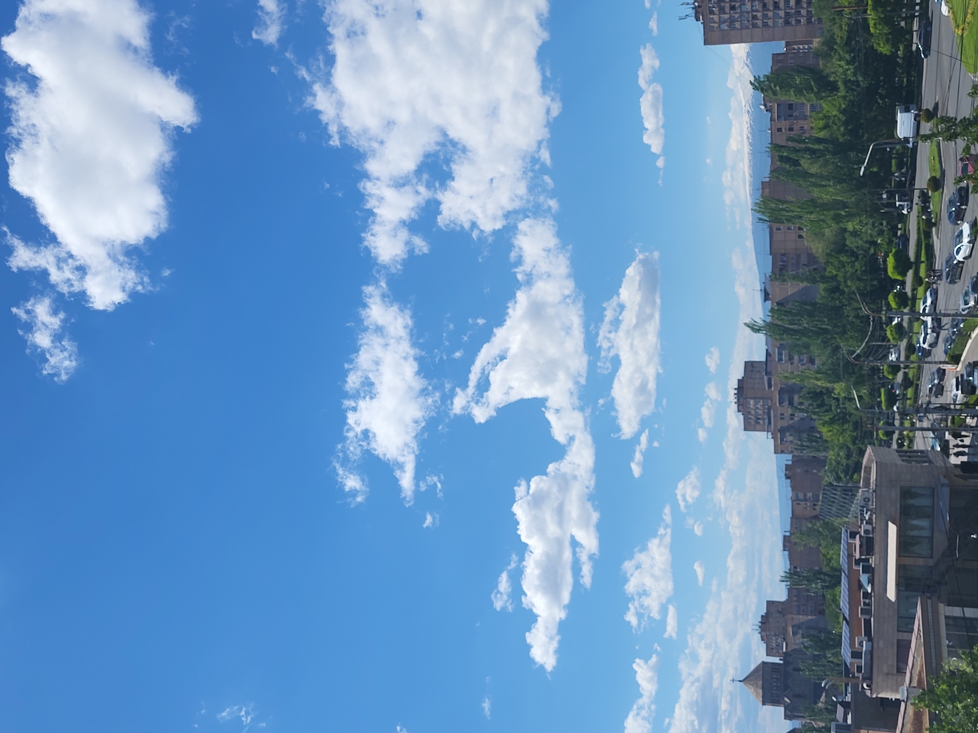 The image is oriented vertically and depicts a cityscape in Yerevan, Armenia, under a bright blue sky scattered with fluffy white clouds. The right side of the frame showcases a street lined with trees and parked or moving vehicles, including cars and possibly a bus. Adjacent to the street are various buildings, ranging from modern high-rises to structures with more traditional architectural styles. In the distance, a hazy, mountainous backdrop can be discerned. The sky occupies the majority of the frame.