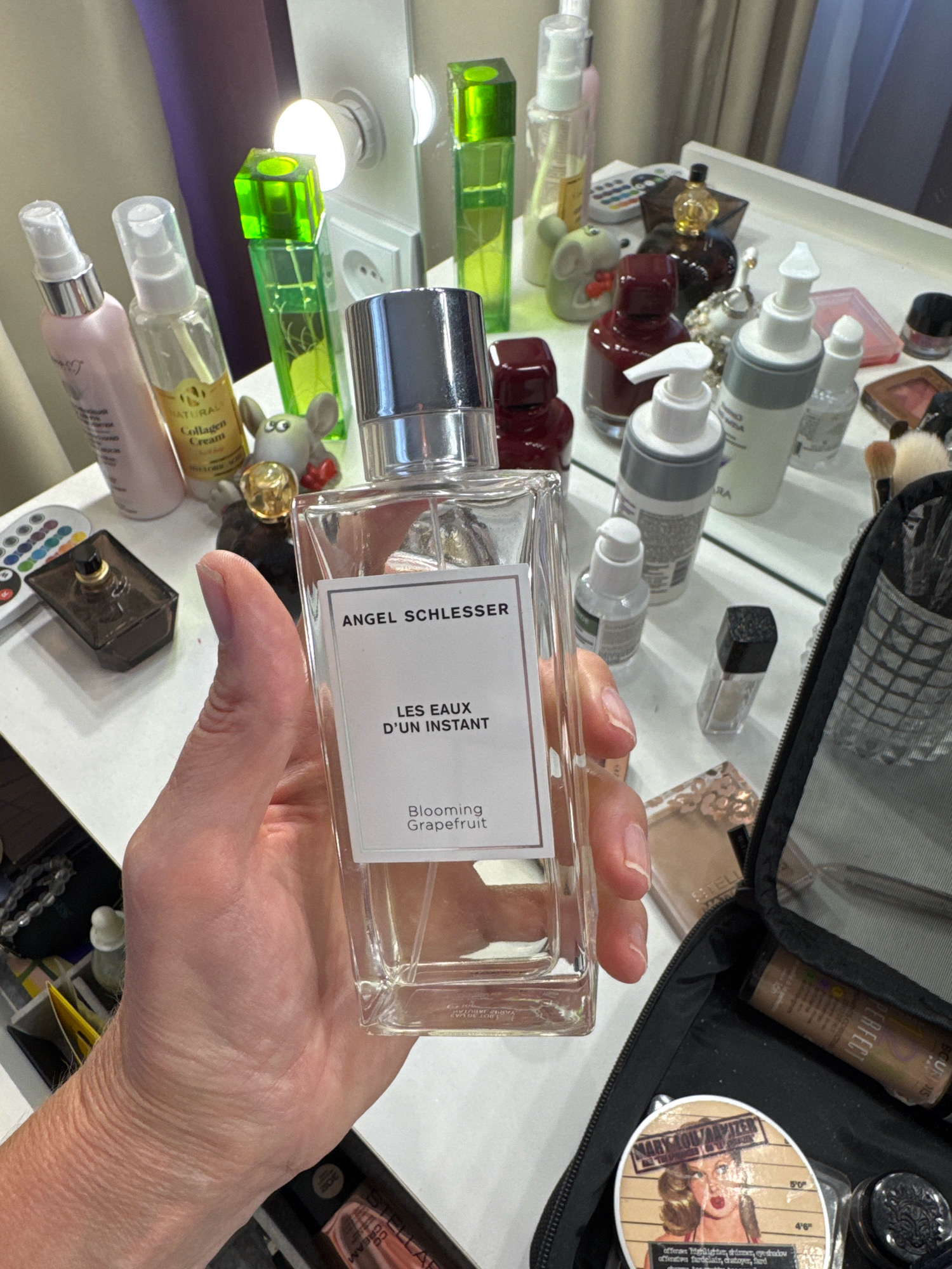 A person's hand holds a clear glass perfume bottle with a silver cap. The bottle has a white label that reads "ANGEL SCHLESSER LES EAUX D'UN INSTANT Blooming Grapefruit." The background shows a vanity table with various cosmetic items, including other perfume bottles, makeup, and brushes. The scene appears to be indoors.