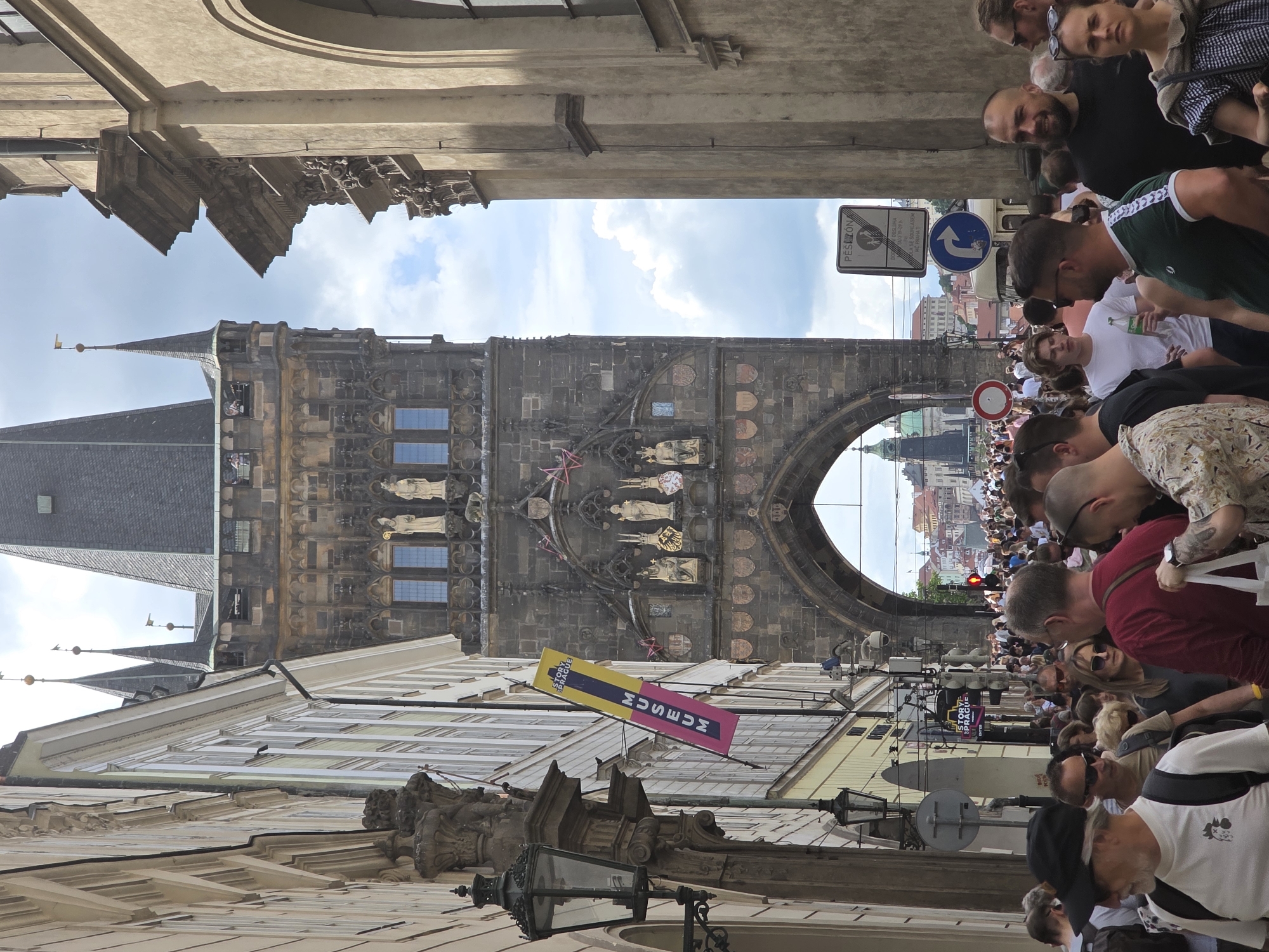 This image depicts a crowded street scene leading towards the Old Town Bridge Tower in Prague, Czech Republic. The perspective is from ground level, looking directly at and through the stone archway of the tower.
In the upper center, the Old Town Bridge Tower dominates, a tall, dark grey Gothic structure with a steep, dark tiled roof. Multiple smaller spires, each topped with a finial and a small flag, adorn the tower. Below the roofline, windows are visible, and the facade is intricately carved with decorative elements, statues in niches, and various coats of arms. The large, rounded archway at the tower's base serves as a passageway. Through this arch, distant city buildings with red tile roofs and a distinct green-domed tower are visible under a bright, partly cloudy sky.
To the left of the tower, a multi-story building with light-colored facades (cream, pale yellow) displays numerous windows. An ornate black street lamp is attached to the building in the foreground. Further back, a vertical yellow and pink banner hangs, bearing the text "HISTORY OF PRAGUE MUSEUM" and "ENTER."
To the right of the tower, a tall, plain tan-colored building with classical architectural details, including cornices and arched windows, rises. Utility poles supporting various signs are present; a blue circular sign with a white right-pointing arrow, a white rectangular sign displaying "PĚŠÍ ZÓNA" (pedestrian zone), and a red circular "no entry" sign are visible amidst the crowd.
The lower half of the image is densely packed with a diverse group of pedestrians, mainly adults, moving towards and through the archway. They are dressed in casual clothing suitable for warm weather, with many wearing sunglasses and carrying backpacks or bags. In the immediate foreground, a man with a white beard and black baseball cap, wearing a white t-shirt with a chicken graphic, walks left of center. To his right, a man in a red t-shirt holds a white tote bag. Further right, a man with a dark beard and black t-shirt smiles slightly, with a woman in a plaid shirt beside him. The scene is brightly illuminated by natural light, casting some shadows.