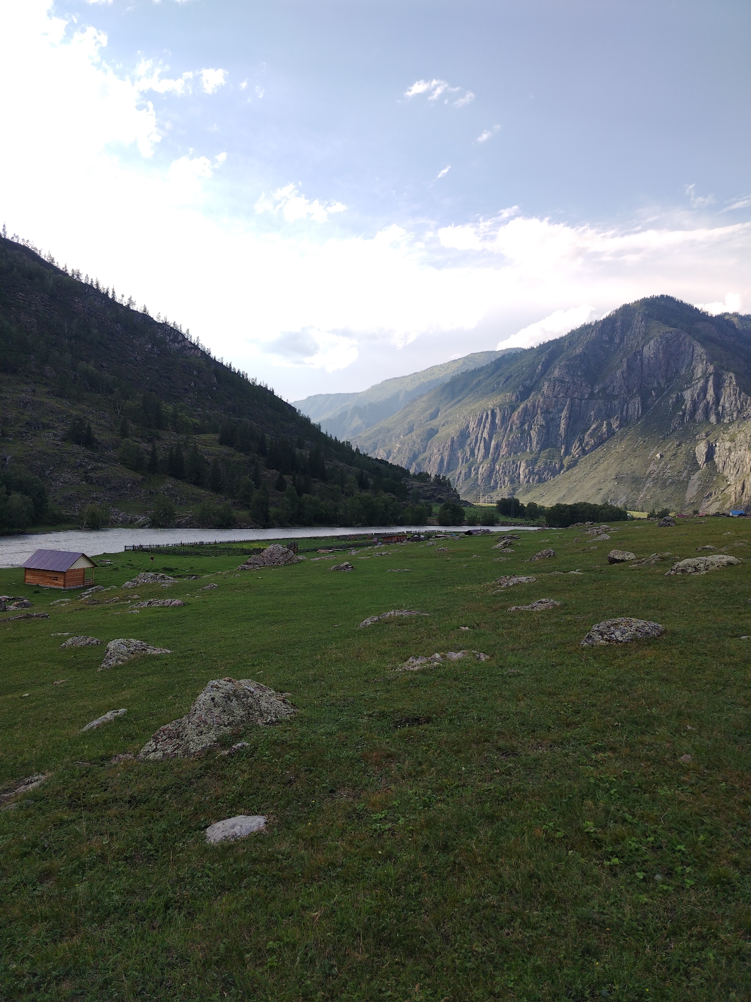 An outdoor, eye-level, medium shot captures a vast, verdant meadow dotted with rocks in the foreground. A meandering river flows in the mid-ground, bordered by a wooden fence. On the left bank of the river, a small, wooden cabin with a dark roof is visible. The background features steep, rocky mountains and a cloudy sky with patches of sunlight. The scene appears to be located in the city of Iodro, Russia. No people are present in the image, and no actions or interactions are depicted.