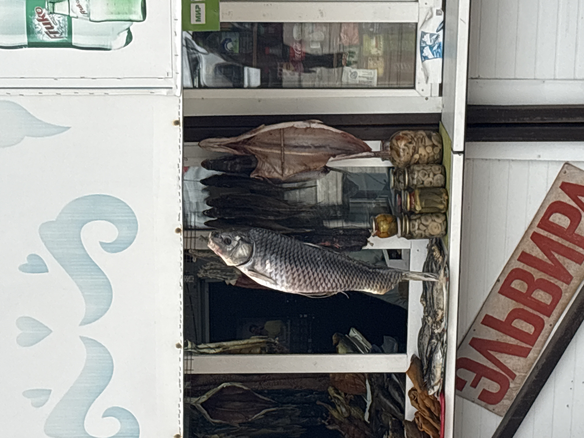 This image captures a display case from a market or shop in Nizhnie Yaki, Russia, showcasing a variety of dried fish and preserved goods. The scene appears to be an outdoor or semi-outdoor retail display, brightly lit during the daytime.
At the center, a large, scaled fish, likely a carp or similar freshwater species, is prominently displayed, hanging horizontally. Its silvery scales and gaping mouth are clearly visible. Below this large fish, several bundles of smaller, darker dried fish are stacked, while above it, a larger, flatter, light-colored dried fish is visible. To the right, additional bundles of dried fish are displayed alongside stacks of small jars or cans that appear to contain preserved items, possibly mushrooms or small pickled foods.
The items are arranged within a white display structure with glass panes. Reflections on the glass reveal shelves inside a shop, holding various packaged products, including what looks like bottled beverages and other retail items. The white panel on the left features light blue decorative swirls and heart shapes. In the upper left corner, two green bottles are partially visible, one bearing the text "Puko." A small green label with the Cyrillic word "МИР" (MIR) is also seen near the top. An angled red and white sign in the bottom right corner displays partial Cyrillic text, reading "ЭЛЬВИР" (ELVIR), which likely indicates a shop or brand name. No people are present in the frame.