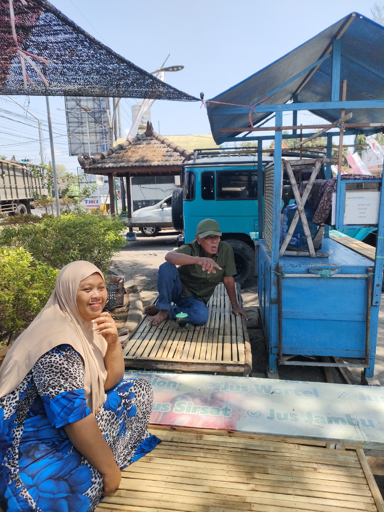 In the city of Curahdringu, Indonesia, a woman wearing a tan hijab and a blue and leopard print dress sits on a bamboo mat. Her left arm rests on her lap and her right hand touches her chin. To her right, a man in a green shirt and cap kneels on a bamboo mat, gesturing with his left hand. Behind them, a blue cart with a blue tarp roof is parked. A turquoise vehicle is partially visible behind the cart. The environment is outdoors, with a clear sky and some vegetation visible. A sign on the cart reads "Daftar Menu" and lists various drink names.