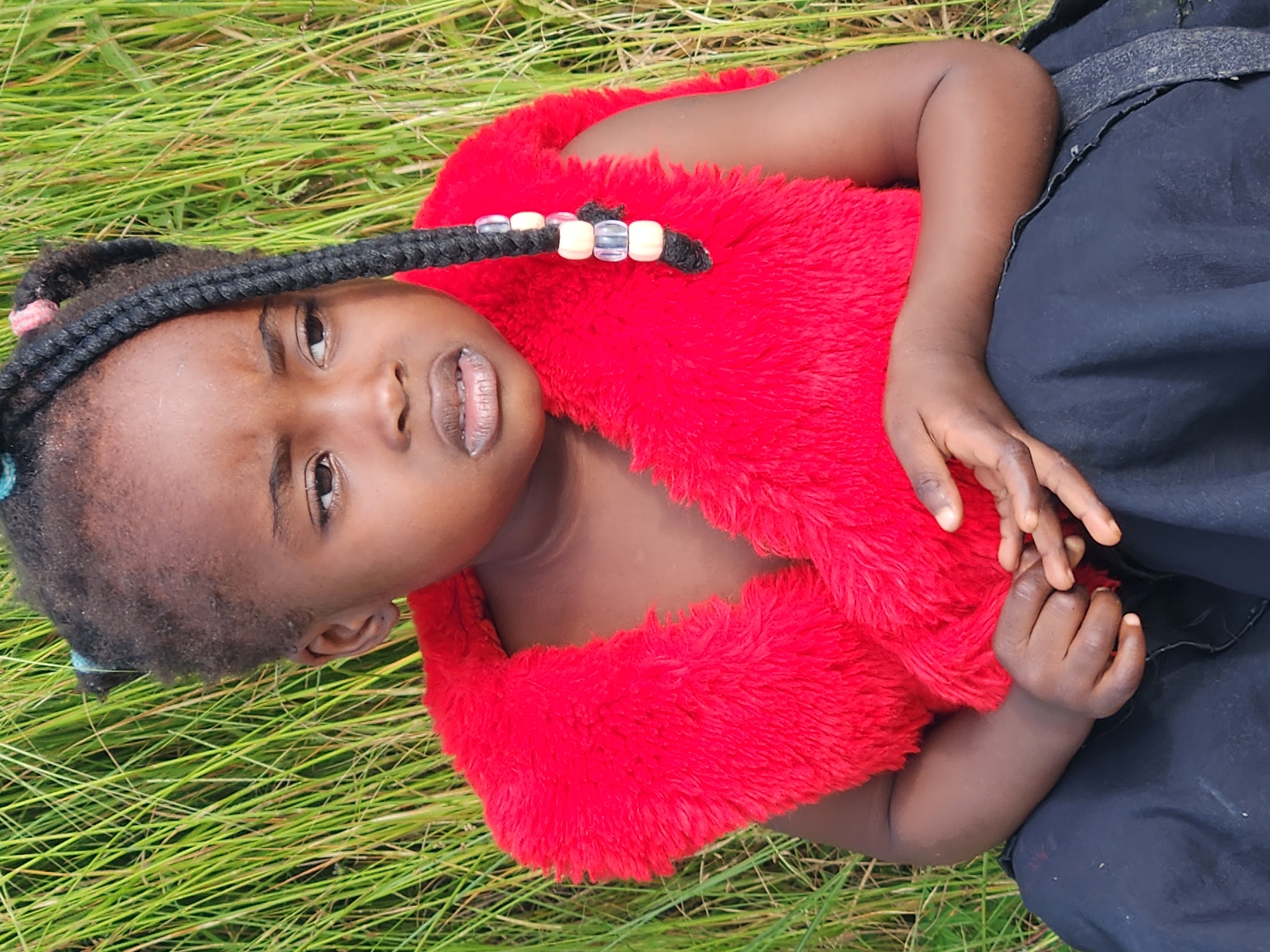 A young girl with dark skin and braided hair adorned with beads sits in front of a backdrop of tall green grass. She is wearing a bright red, furry vest over a dark garment. Her hands are clasped in her lap, and her gaze is directed away from the camera with a thoughtful expression. The lighting suggests an outdoor daytime setting. This image could have been taken in the city of Jos, Nigeria, a region known for its verdant landscapes and vibrant culture.