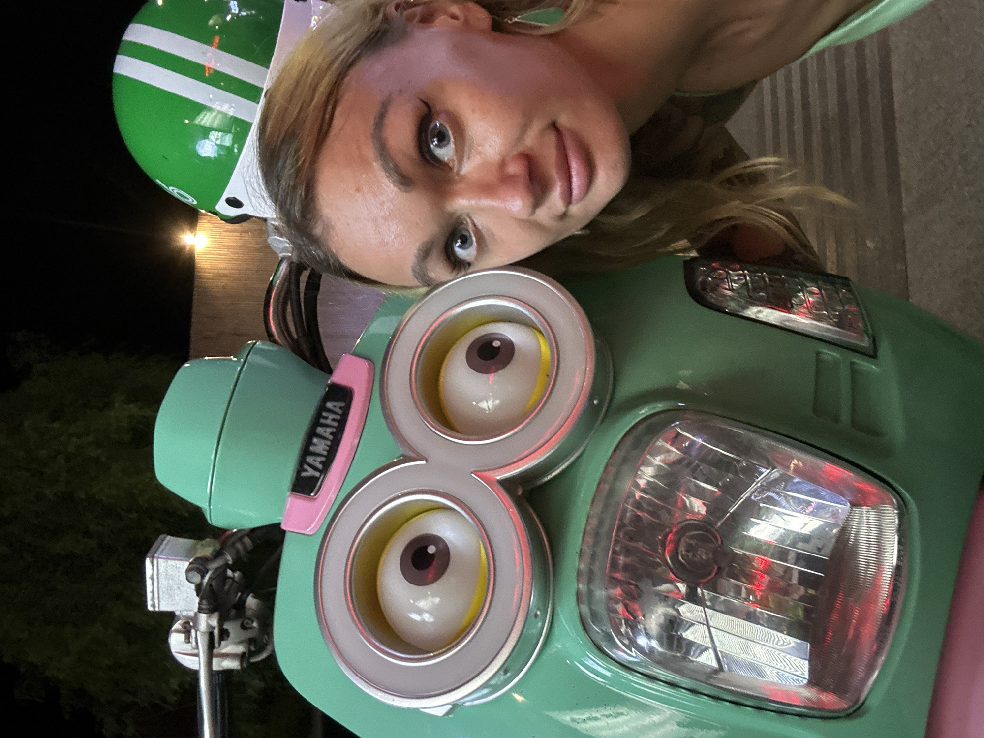 A medium shot image captured at night shows a blonde woman posing for a selfie next to a teal-colored scooter that has been modified to resemble a Minion character from the animated film franchise. The scooter's front features two large, circular headlights that mimic the eyes of a Minion, complete with white irises, brown pupils, and yellow accents. A pink band with the "YAMAHA" logo is affixed above the Minion-like eyes. The scooter also has a bright green helmet with white stripes on top, positioned to the right of the Minion eyes.
The woman, who is positioned on the right side of the frame, looks directly at the camera with her eyes open and a slight smile. She is wearing a light green top and has on noticeable eyeliner and green teardrop-shaped earrings. Her blonde hair is partially obscuring her right shoulder.
The background is dimly lit, suggesting an outdoor setting at night. A wooden fence and some foliage are visible behind the scooter and the woman. Streetlights are casting a warm glow on the scene. The scooter's headlights and taillights are illuminated, reflecting red and white light. The pavement or ground beneath them appears to be textured. The overall impression is that this scene took place in an urban environment, possibly in Phu Thuy Ward, Vietnam, given the context.