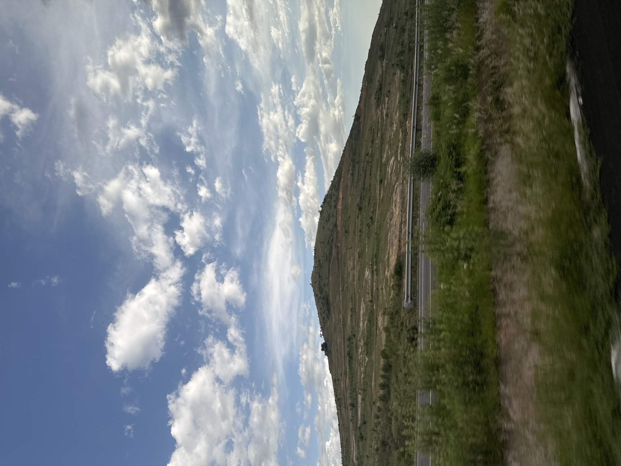 The image depicts a natural landscape dominated by a large, grassy hill under a partly cloudy blue sky. The hill exhibits terraced formations, suggesting agricultural or land management practices, with patches of bare earth and sparse vegetation. A highway runs horizontally across the midground, separated from the viewer by a barrier and lush green foliage. The foreground is filled with blurred green vegetation, indicating movement or a shallow depth of field. Several trees are visible on the crest of the hill. The overall scene conveys a rural or semi-rural environment.