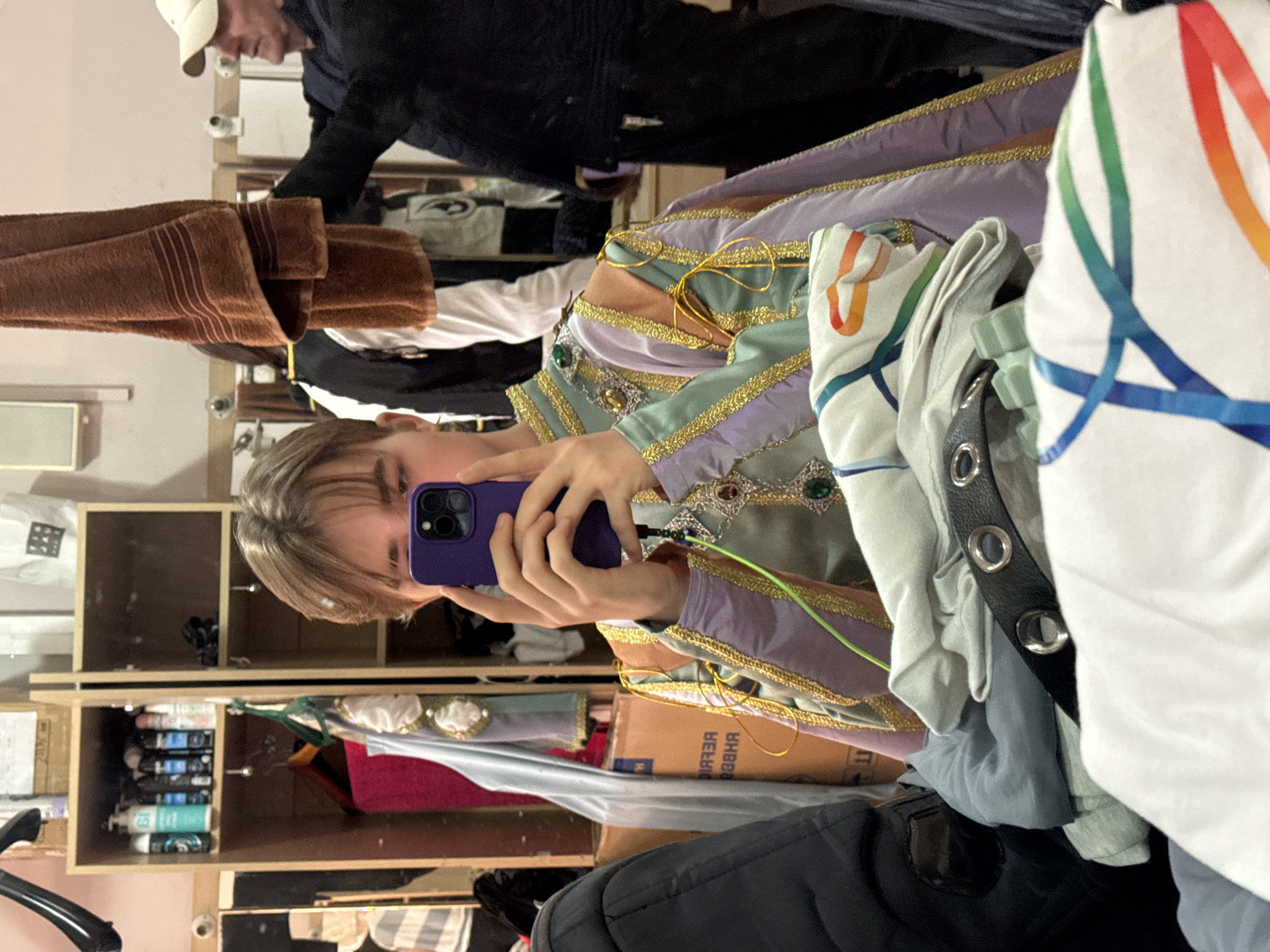 A person is taking a selfie in what appears to be a cluttered backstage or dressing room. The person is holding a purple smartphone and is wearing an elaborate costume with green, gold, and lavender detailing and possibly some decorative jewels. The background reveals shelves containing various items, including hair products, a box, and clothing. Another person wearing a baseball cap and dark clothing is visible in the background. The environment suggests an indoor setting, likely associated with a theatre or performance venue. No specific details indicate the city of Tbilisi, Georgia.