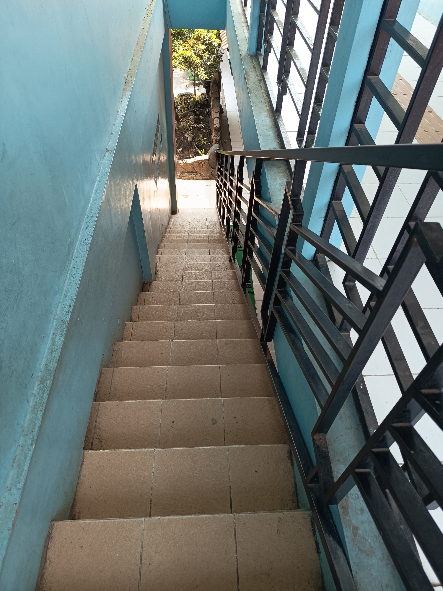 The image shows a downward view of a tiled outdoor staircase in Widoro, Indonesia. The staircase has a light brown tiled surface and is flanked by a pale blue wall on the left and a black metal railing on the right. The railing has a geometric pattern with horizontal and diagonal bars. Beyond the railing, a white tiled surface is visible. At the top of the stairs, there is an opening that looks out onto a small garden area with trees and plants, illuminated by bright sunlight. The overall scene suggests an exterior stairway likely part of a building or residential area.