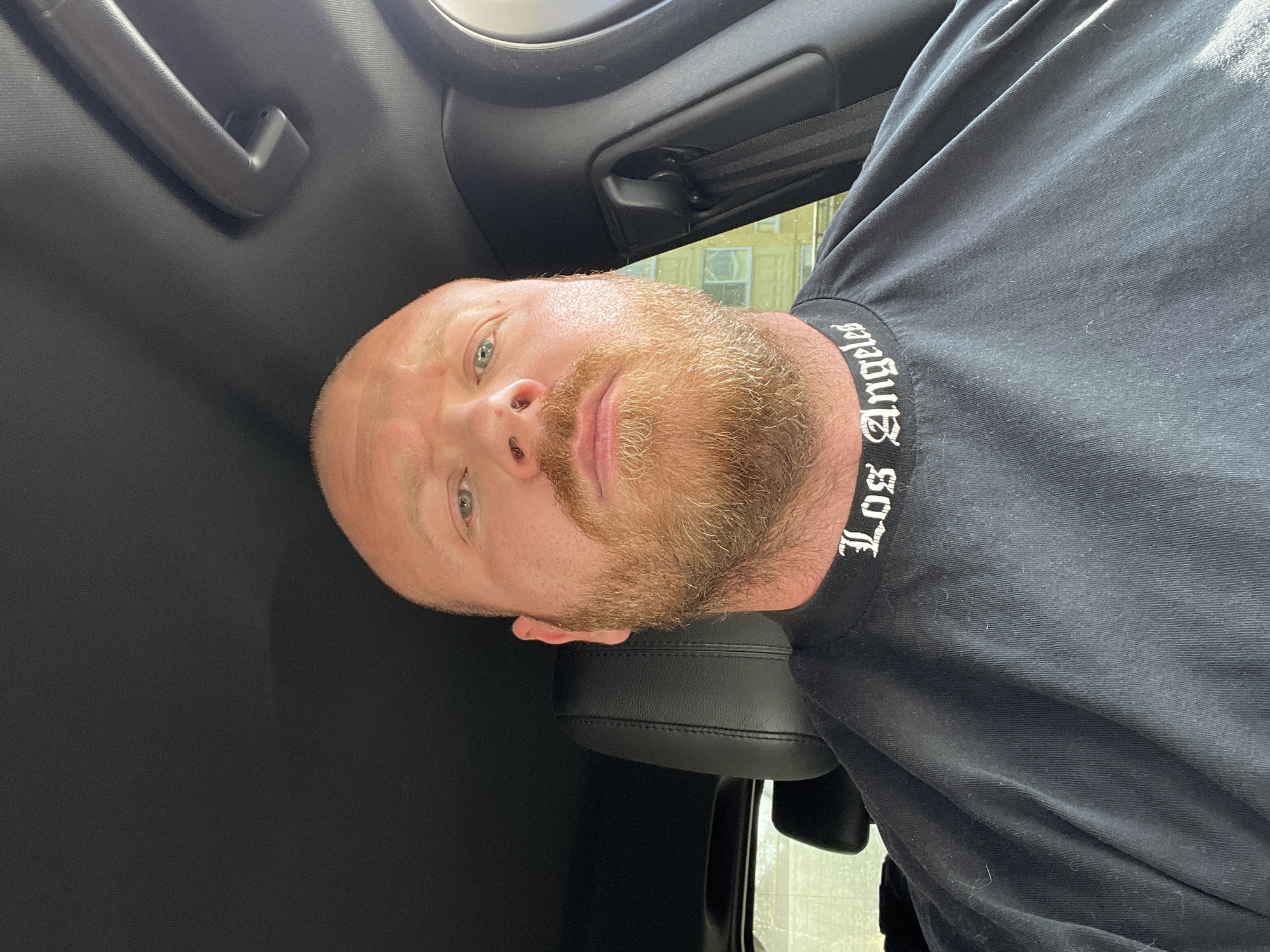 A man with fair skin, a bald head, a ginger beard and blue eyes is seated in the driver's seat of a car. He is wearing a black t-shirt that reads "Los Angeles" in a white gothic font. The interior of the car is black, and a grab handle is visible on the ceiling. The location is unknown.