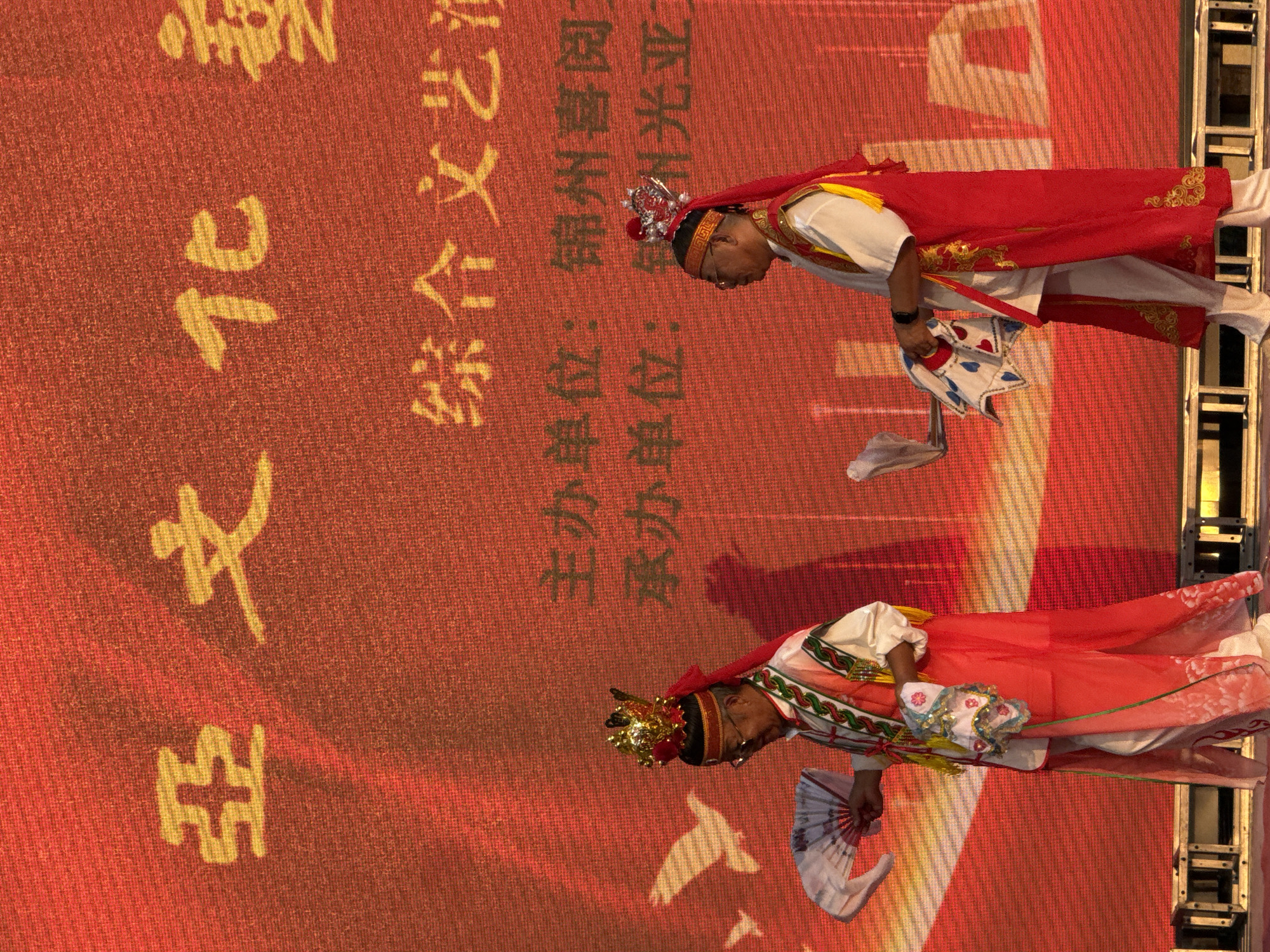 The image depicts a stage performance with two individuals dressed in traditional Chinese attire against a large red backdrop featuring yellow Chinese characters. The characters at the top appear to read "亞文化" which translates to "Asian Culture." Below this, additional text is visible, including "綜合文藝" (Comprehensive Arts) and "主辦單位: 錦州喜閱" (Organizer: Jinzhou Xiyue) and "承辦單位: 錦州光亞" (Undertaker: Jinzhou Guangya). The individuals are performing a dance, each holding a patterned fan. The person on the left wears a golden headdress with red ribbons and a red and white costume adorned with colorful embroidery. The person on the right wears a silver headdress with red ribbons and a red vest over a white garment. Both are wearing white pants. The backdrop has a subtle gradient effect, transitioning from orange at the bottom to red at the top, and features faint white silhouettes of birds. The stage has metallic scaffolding visible at the bottom. The scene takes place in Jinzhou, China, indicated by the text on the backdrop.