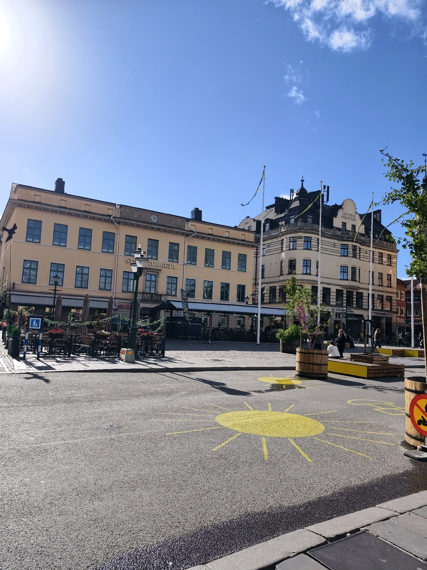 The image shows a street-level view of Stora Torget in Linköping, Sweden, on a sunny day. In the foreground, yellow sun drawings are painted on the asphalt, indicating a playful or decorative element within the public space. Two large wooden planters are situated on the right side of the image, one with a sign depicting a crossed-out bicycle, suggesting a designated no-cycling zone or a children's play area. Beyond the sun drawings, the scene opens up to a paved area with outdoor seating for restaurants and cafes. Several tables and chairs are visible, some covered by umbrellas.
The buildings in the background are of varying architectural styles. The building on the left is a pale yellow structure with multiple windows and a sign that reads "STO(RA) TORGET 1". A lamppost stands in front of it. To the right of this building is a larger, more ornate building with a dark roof featuring spires. This building displays flags and has signage that includes "CENTRAL PALATSET". People are scattered throughout the scene; some are seated at tables, while others are walking or standing. The sky is clear blue with a few wispy clouds, and the sun is bright, casting shadows. The overall impression is of a lively public square on a pleasant day.
