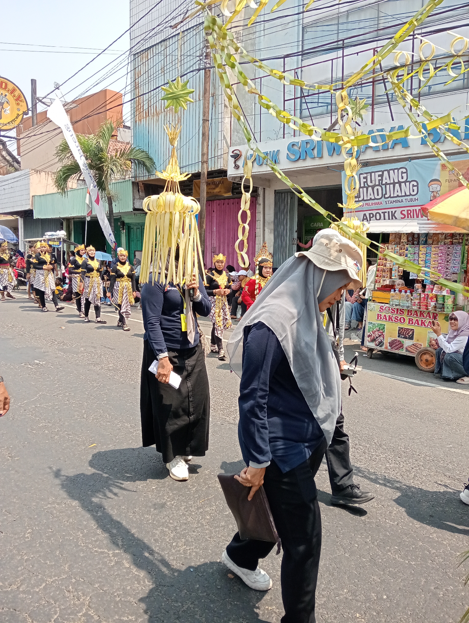 This is a medium-shot, eye-level, outdoor photograph taken on a sunny day, likely in the city of Demak, Indonesia. The image captures a street scene with a procession or parade. In the foreground, a person wearing a dark blue long-sleeved shirt, black pants, and a light gray hijab is walking towards the right of the frame. They are carrying a dark brown clutch bag in their right hand and appear to be looking down. A second person, slightly behind and to the right, is also walking and looking towards the right. They are wearing a dark blue long-sleeved shirt and black pants.

Further into the scene, a group of individuals dressed in traditional costumes are marching in a line. They wear black tops with gold, white, and red patterned vests and skirts, along with gold crowns. Some are carrying what appear to be props. Behind this group, buildings line the street, with some displaying signage that may indicate shops or businesses. Notably, a banner with text in Indonesian, including "FUFANG JIAOJIANG" and "APOTIK SRIWIJAYA," is visible on one of the buildings.

Decorative elements, possibly made from palm leaves, are suspended across the street and from poles, adding to the festive atmosphere. A food stall selling what looks like grilled sausages (sosis bakar) and bakso is also visible to the right. The street surface is asphalt, with some cracks and wear visible. The overall scene suggests a cultural event or celebration taking place in a commercial or urban area of Demak.