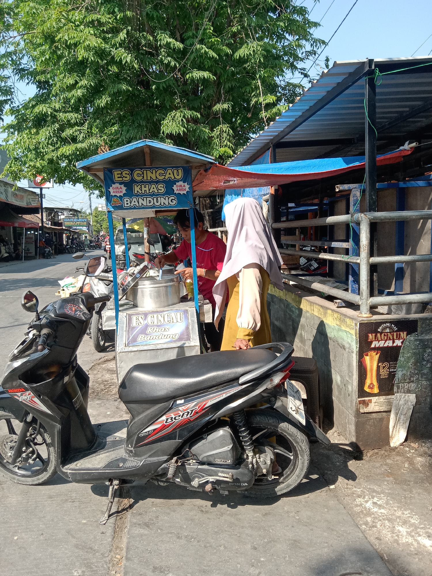 In Sugihwaras, Indonesia, a street scene unfolds under a clear sky and the shade of a large tree. A vendor, wearing a red shirt, stands behind a mobile cart selling "Es Cincau Khas Bandung." The cart has a blue awning and is adorned with text in Indonesian, including "ES CINCAU KHAS BANDUNG." The vendor is actively preparing a drink, using a ladle in a large stainless steel container. Beside the cart, a woman in a light purple hijab and a yellow garment stands, partially obscured. A black Honda Beat scooter is parked in the foreground, angled towards the street. The street itself is paved and shows signs of wear. Beyond the immediate scene, other parked motorcycles, buildings, and electrical wires are visible, suggesting a commercial or residential area. A poster for "MAGNUM MAX" is affixed to a wall on the right side of the image. The overall atmosphere is that of a sunny day in a bustling Indonesian town.