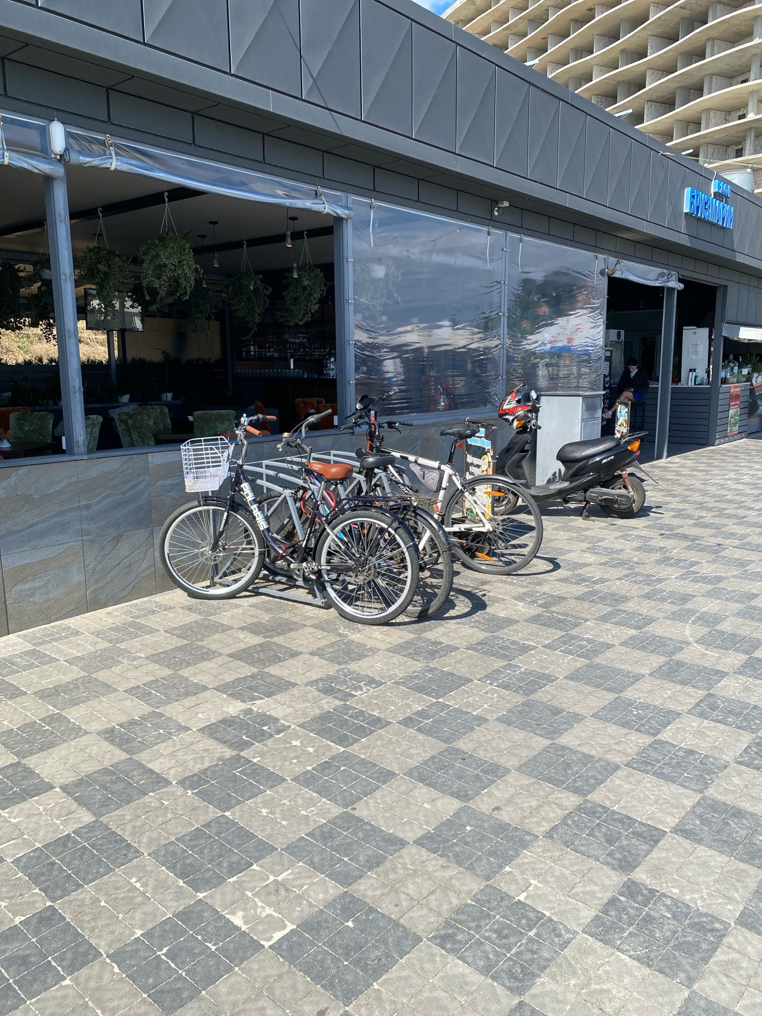 The image captures a sunny day scene outside a building with a transparent plastic curtain enclosure. Several bicycles are parked to the left, with one prominently featuring a white basket. To the right of the bicycles, a black scooter is parked. A person wearing a black top and blue shorts is seated on a chair inside the establishment, visible through an open doorway. The building's facade is modern, with a grey metallic finish. Above the establishment, a large unfinished concrete building is visible against a bright blue sky. The ground is paved with a checkered pattern of grey and light-colored square tiles. The location is identified as Odesa, Ukraine.