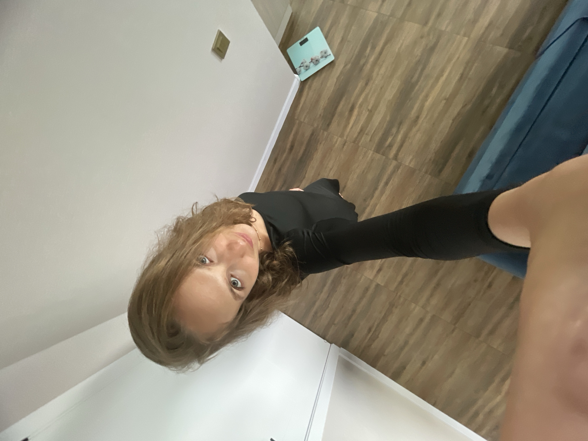 This is a selfie taken by a person with shoulder-length brown hair and blue eyes. They are wearing a black long-sleeved shirt and black leggings. The person is standing indoors, and the scene includes a white wall, light brown wooden flooring, and a blue couch or chair. In the background, there is a light blue scale with a floral pattern on it. A light switch is visible on the white wall. The lighting suggests it is daytime. The person is looking directly at the camera with a neutral expression. The overall setting appears to be a residential interior, possibly a bedroom or hallway.