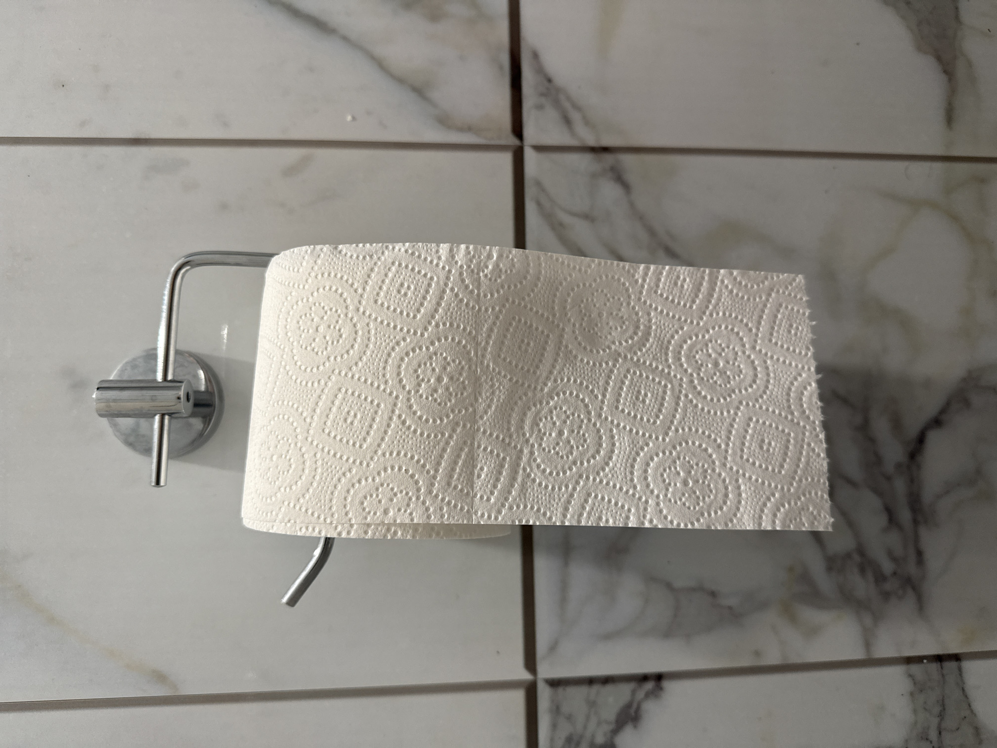 A roll of white toilet paper hangs on a chrome holder mounted on a wall tiled with white and gray marble-patterned tiles. The toilet paper has a textured pattern of circles within squares. The lighting is overhead, casting slight shadows. The scene is indoors, likely a bathroom.