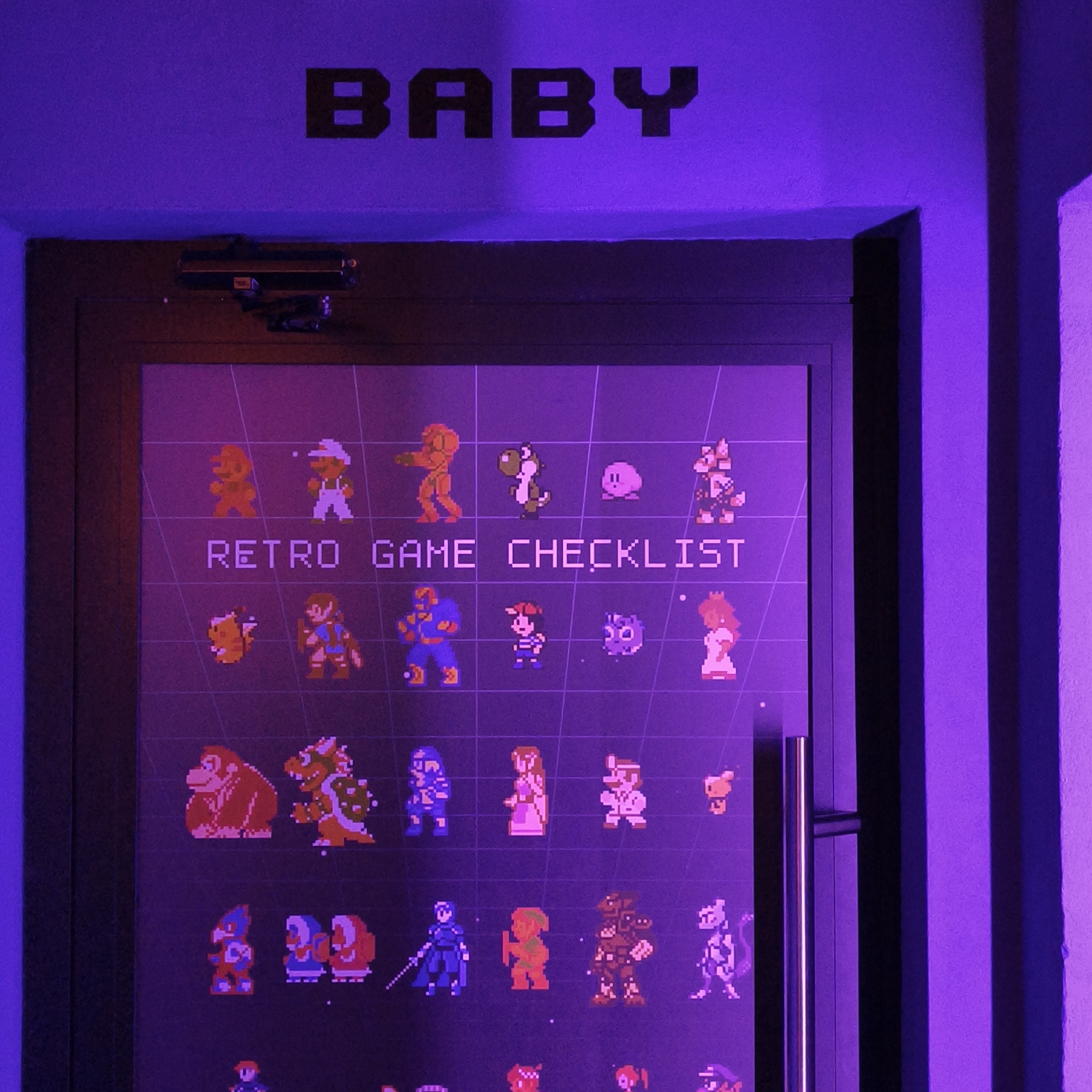 A color photograph taken indoors under purple lighting, displays a dark door with a metal handle, bearing a framed retro game checklist. The word "BABY" is stenciled in black above the door. The checklist features pixelated character sprites from various retro video games arranged in rows and columns, against a dark background overlaid with a grid pattern. The door has a closer mechanism attached at the top. The interior wall is painted in a similar shade of purple. The photograph does not provide enough information to determine its specific location in Hrodna, Belarus. No people or interactions are visible in the image.