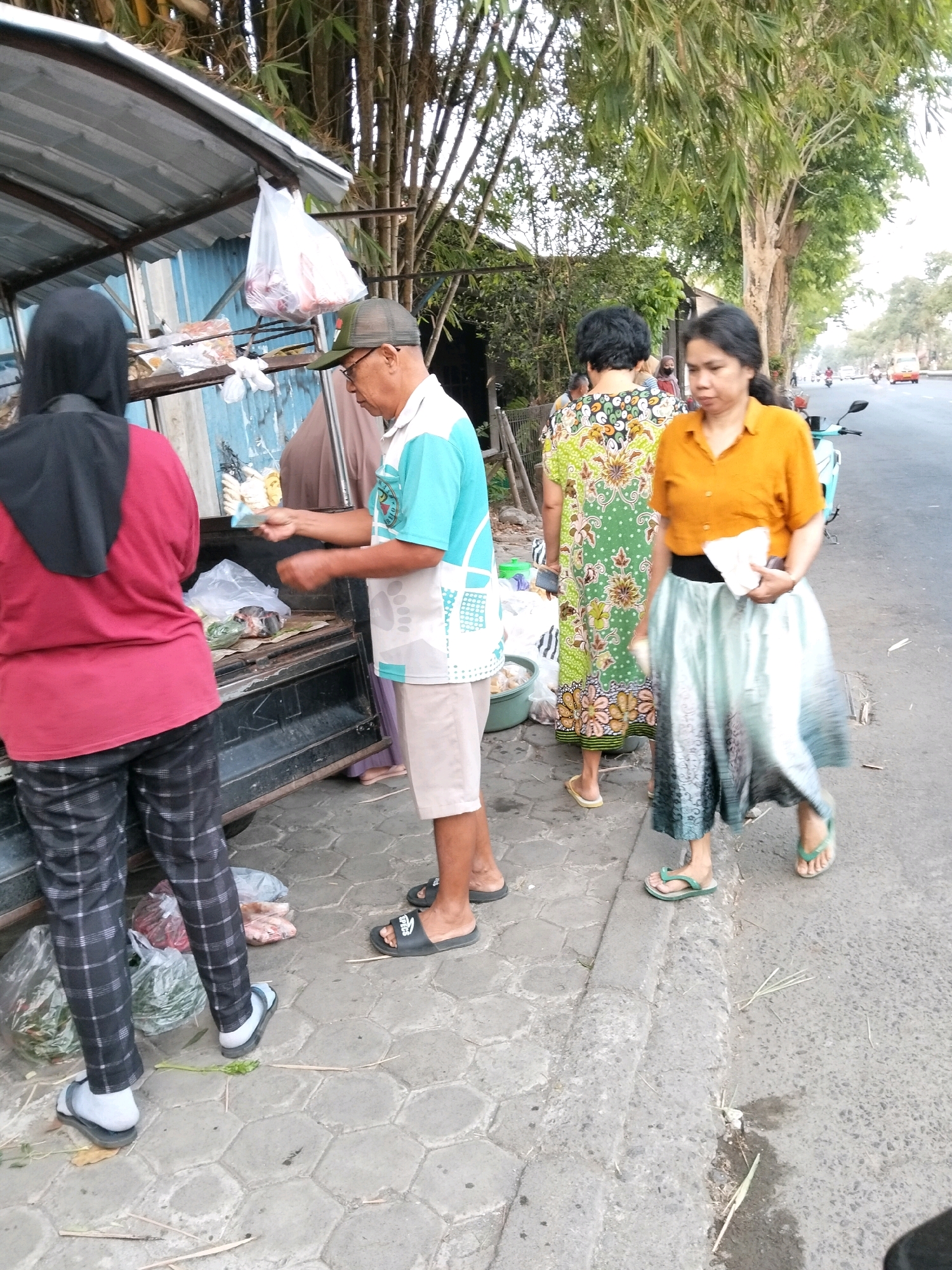 In Pangenjurutengah, Indonesia, a street vendor sells goods from a makeshift stall. A woman wearing a red shirt and black headscarf stands at the stall, looking at the goods. Next to her, a man in a blue and white shirt and khaki shorts appears to be making a transaction with the vendor. Another woman in a green and white patterned dress stands behind them, while a third woman in an orange shirt and a flowing skirt walks past. The stall itself is a flatbed truck with a metal roof, displaying various items for sale, including what appears to be meat wrapped in plastic. The scene is set on a paved sidewalk next to a road, with a line of trees providing shade and a glimpse of traffic in the distance.