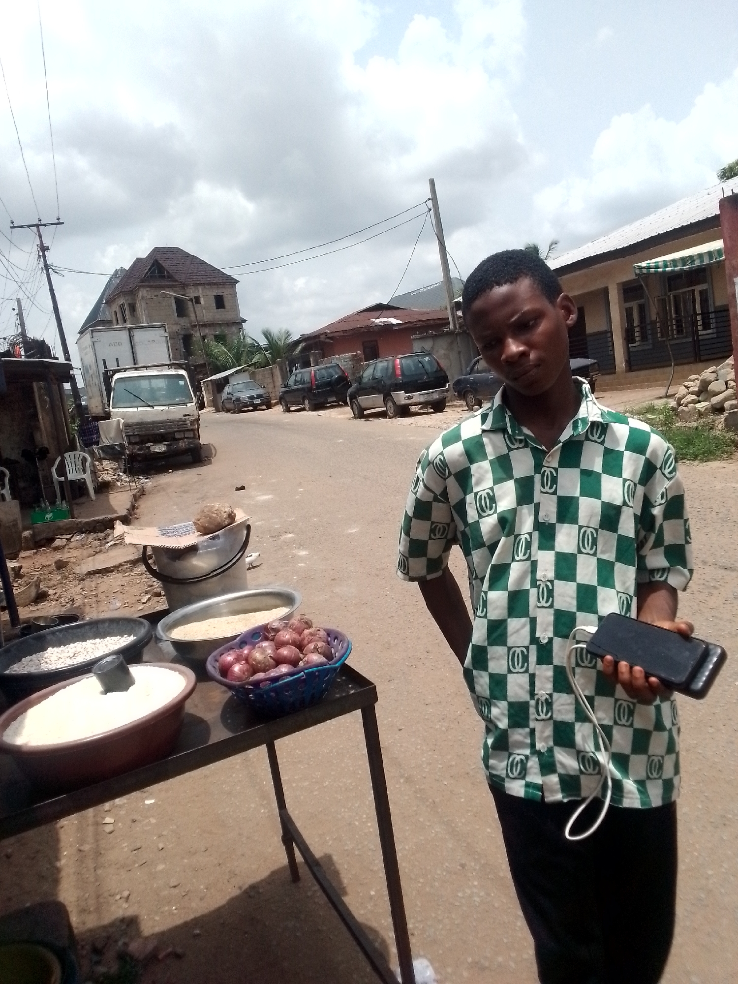 A young man stands on an unpaved street in Onitsha, Nigeria. He is wearing a green and white checkered shirt and black pants. He holds a black smartphone connected to a white charging cord in his right hand.
Behind him to the left, a dark metal table holds several containers. One contains white grains, another contains onions, and another contains unidentified white food. To the left and behind the table, there is a parked white truck, a building under construction, and a street with parked cars.
The sky is overcast with grey clouds. Power lines are visible overhead. A building with an awning is visible on the right side of the frame.