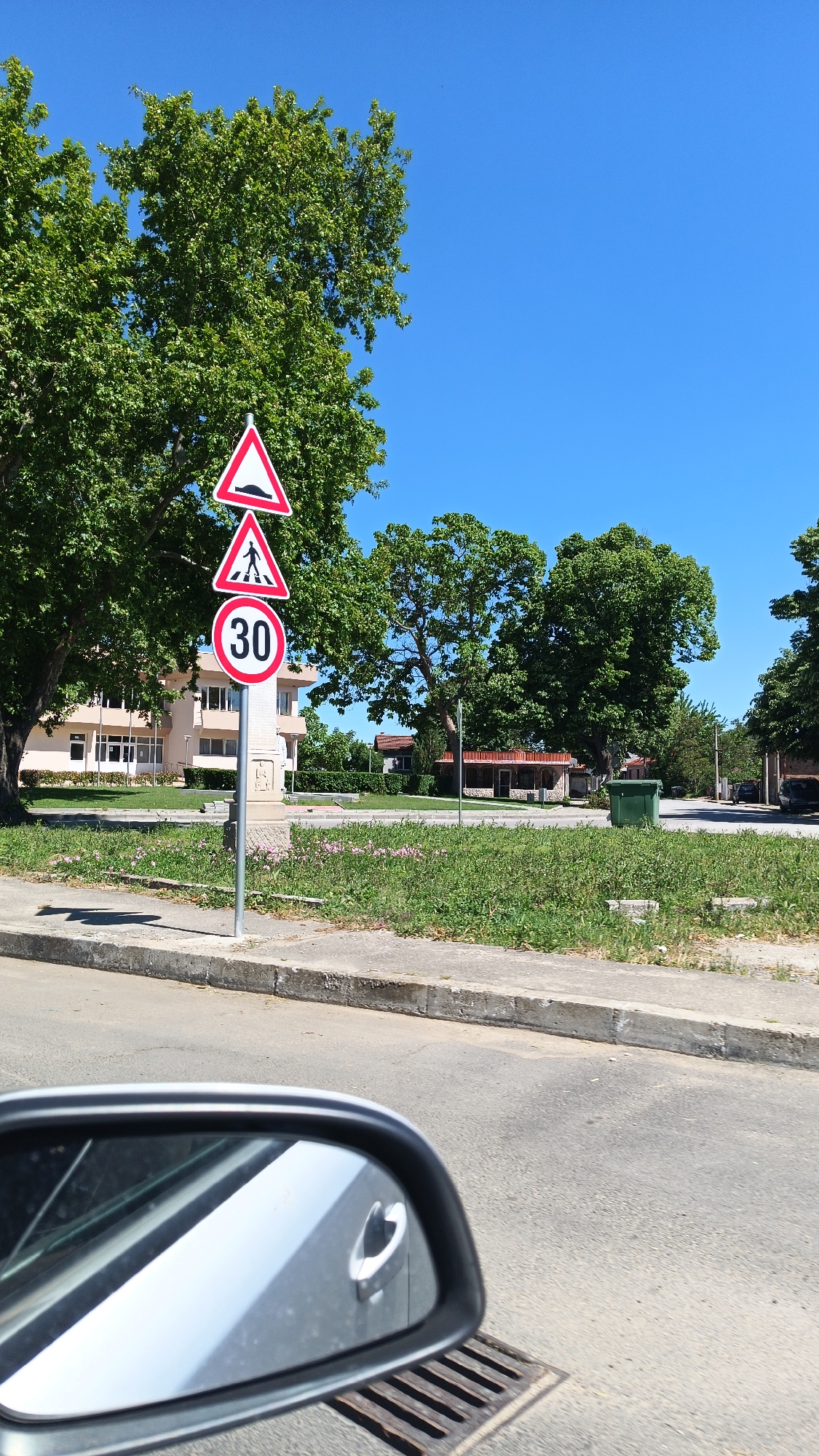 The image shows a street scene in what appears to be Milevo, Bulgaria. A road sign is prominently visible, displaying three warnings: a bump ahead, a pedestrian crossing, and a speed limit of 30. The sign is positioned on a pole next to a sidewalk with grass and pink flowers growing on the other side. A beige building with windows is visible in the background, along with trees and other structures. A green trash bin stands near a small structure with a red roof. The view is from inside a car, with the car's side mirror partially visible in the foreground, reflecting a gray or silver exterior. A grate or drain is also visible near the mirror. The sky is clear and blue.