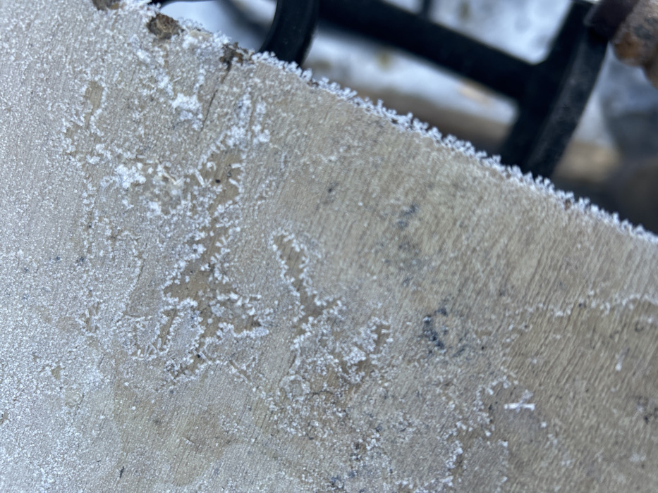 The image displays a close-up, oblique view of a weathered wooden surface, extensively covered with crystalline frost formations, in Chaikovsky, Russia. The wood exhibits visible grain patterns and areas of minor discoloration. The frost is particularly dense and jagged along the exposed right edge of the wooden object, forming a distinct white fringe. Irregular, branching frost patterns, resembling delicate dendritic structures, are distributed across the surface. In the blurred background, elements consistent with an outdoor, freezing environment are discernible, including a snow-covered ground surface and dark, rectilinear components of what appears to be a metallic structure or furniture support. No people, specific actions, or interactions are visible.