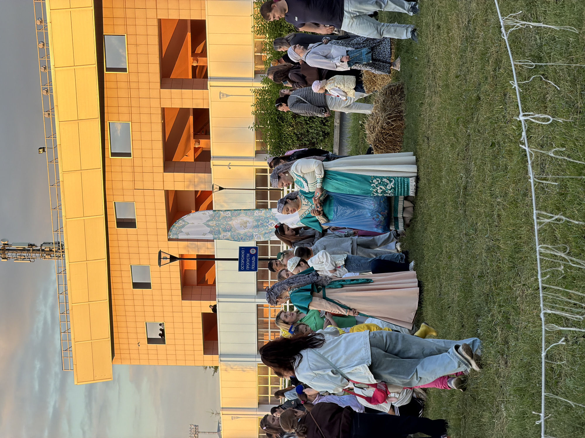 In the city of Unknown, Unknown, an outdoor gathering is taking place in front of a modern, orange-tiled building with a yellow upper structure housing a large antenna. The building has multiple rectangular windows and open-air sections. A group of people are milling about on a grassy area scattered with hay bales. Several individuals are wearing traditional clothing, including a woman in a long, teal and white embroidered dress with a headscarf, and another woman in a similar, lighter colored dress. Other attendees are dressed in casual attire. A blue sign with Cyrillic text is visible, appearing to be a traffic or event marker. The lighting suggests it is late afternoon or early evening, with warm, golden sunlight illuminating the scene. The foreground is partially obscured by a string barrier.