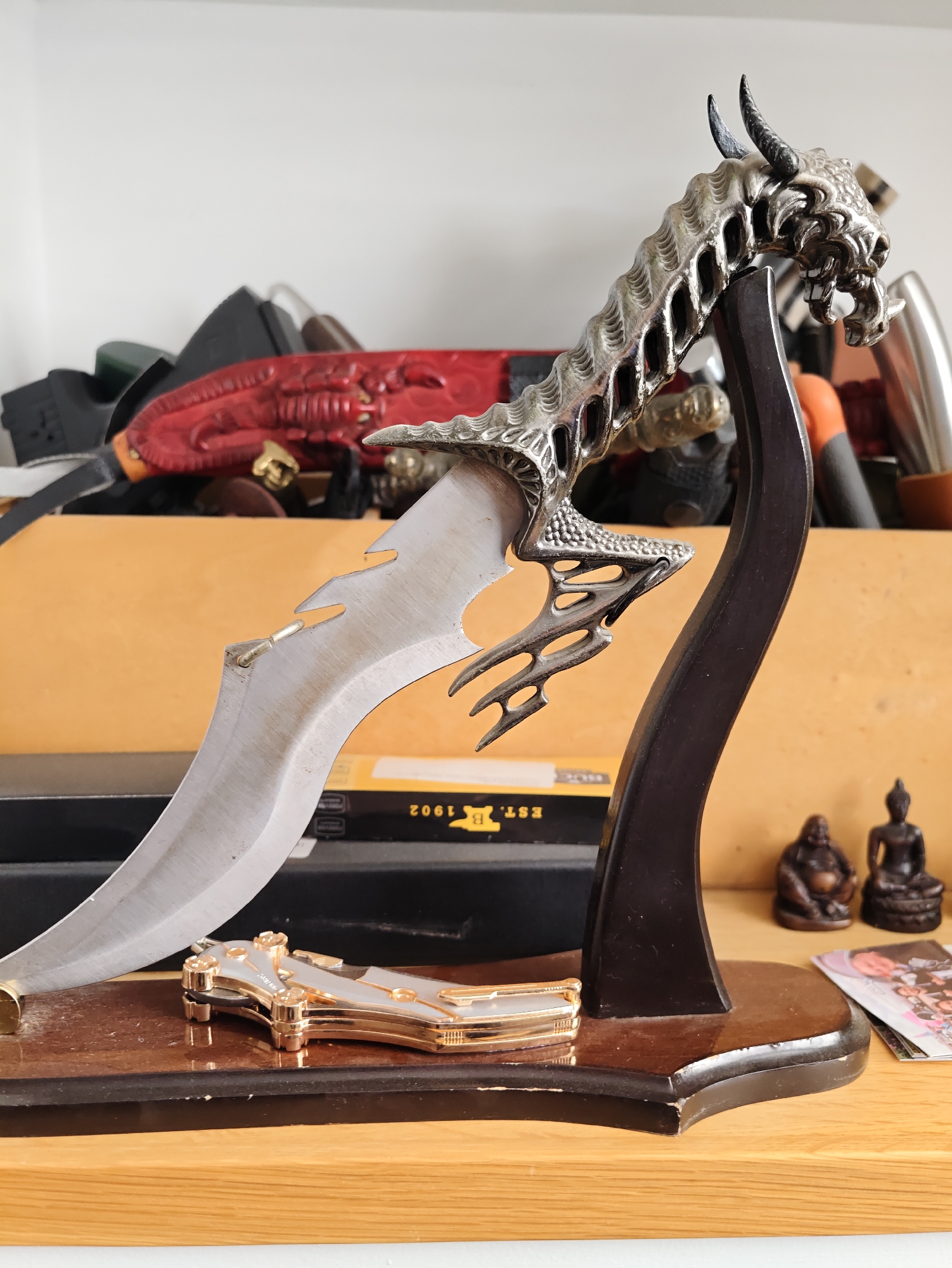 The image displays a collection of knives on a wooden shelf. In the foreground, a decorative knife with a dragon-shaped hilt is prominently displayed on a dark wooden stand. The dragon hilt is intricately detailed, with scales, horns, and a fierce expression. The blade of this knife is curved and has a serrated edge. To the left of the dragon knife's stand, a smaller, ornate pocket knife with gold-colored accents is visible.
In the background, several other knives are partially visible, some with red, black, or silver handles. Two small Buddha statues are placed to the right of the main display, along with what appears to be a stack of cards or photographs. The shelves appear to be made of wood, and the overall setting suggests a personal collection or display. The image was taken indoors, with a plain white wall in the upper portion of the frame. The lighting is natural and evenly distributed. No people are present in the image, and no specific location in Petah Tikva, Israel, can be identified from the visual information provided.