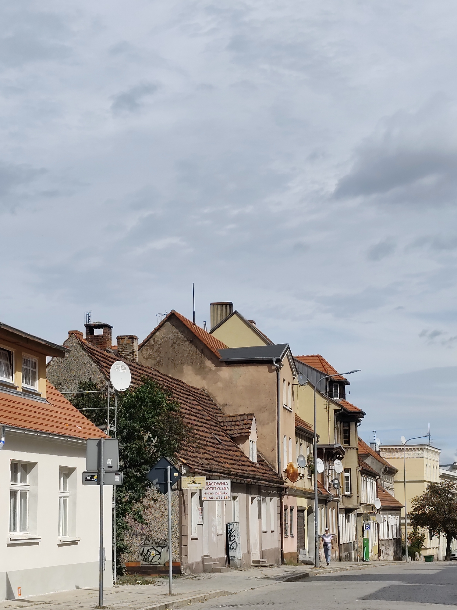 The image shows a street scene in Zielona Góra, Poland, with a row of attached buildings on the right and a white building on the left. The buildings have red tile roofs and are of varying architectural styles and ages. There are satellite dishes attached to some of the buildings. A person wearing a striped shirt and dark pants is walking on the sidewalk. There are street signs and some graffiti on the buildings. The sky is overcast with clouds.