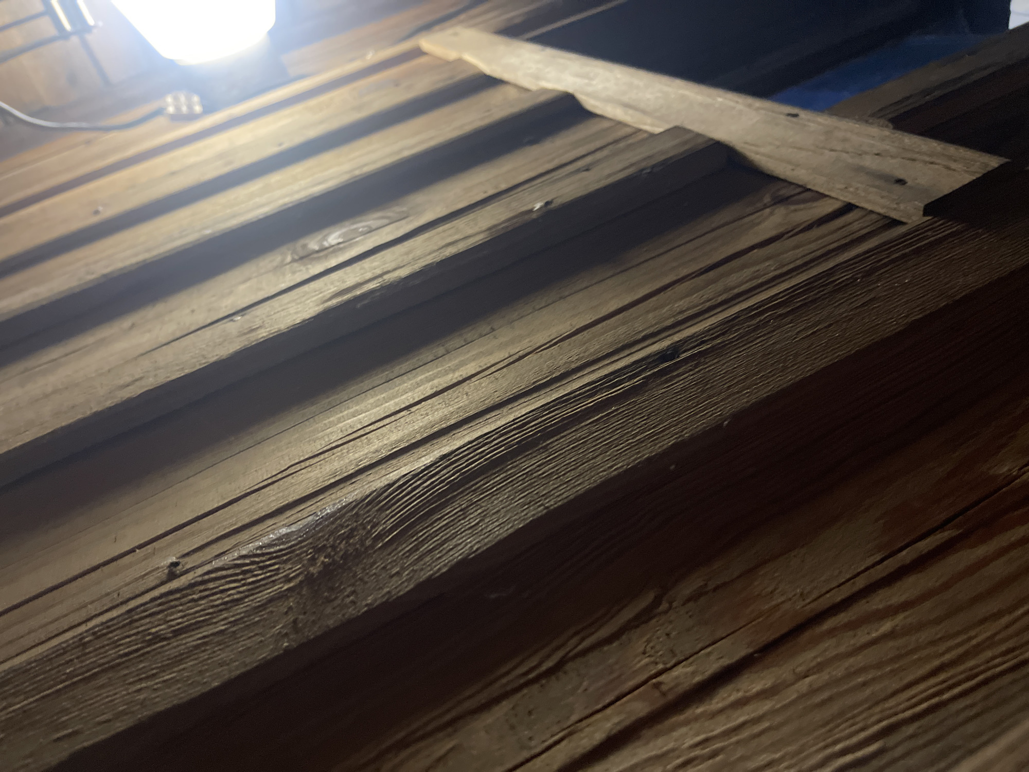 The image shows a close-up, angled view of weathered wooden planks, likely part of an attic or interior wall. The planks are oriented diagonally and appear to be constructed of rough-hewn wood with visible grain and some discoloration. A thin, lighter-colored wooden piece is attached diagonally across some of the planks in the lower right portion of the frame. A bright light source, possibly a bulb, is visible in the upper right corner, casting light and shadows on the wooden surfaces. No people or specific environmental context outside of the wooden structure are discernible. The location is identified as Sibirskiy, Russia.