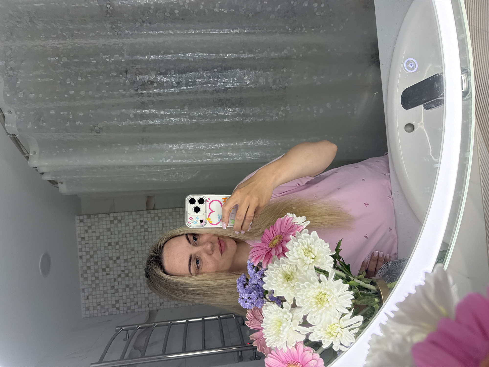 A woman with blonde hair holds a bouquet of white, pink, and purple flowers in front of her as she takes a selfie in a bathroom mirror. She wears a pink shirt. The bathroom features a shower curtain, patterned wall tiles, and a white sink. The location may be Votkinsk, Russia.