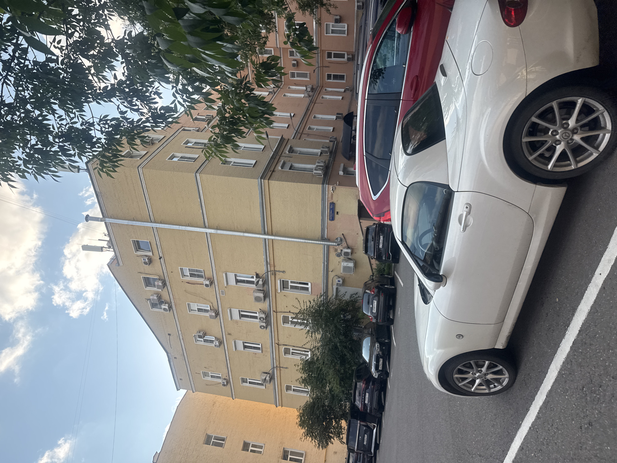 The image depicts a daytime exterior scene in Moscow, Russia, focused on a parking lot in front of a building complex. In the foreground, a white convertible sports car is parked, partially obscuring a red SUV and several other dark-colored cars parked behind it. The buildings in the background are multi-story structures with yellow and orange facades, featuring numerous windows and external air conditioning units. Trees with dense green foliage are visible, framing the scene on the right side and between the buildings. The sky is partly cloudy, with a few wispy clouds against a blue backdrop. A street sign with Cyrillic lettering is affixed to the building on the left, indicating the location as "МАЛЕНЬКАЯ БРОННАЯ / 14/16".
